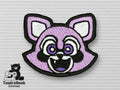 Embroidered patch of a stylized, smiling purple raccoon face with white and black details, displayed on a textured light gray surface with the TundraGeek Creations logo in the corner.