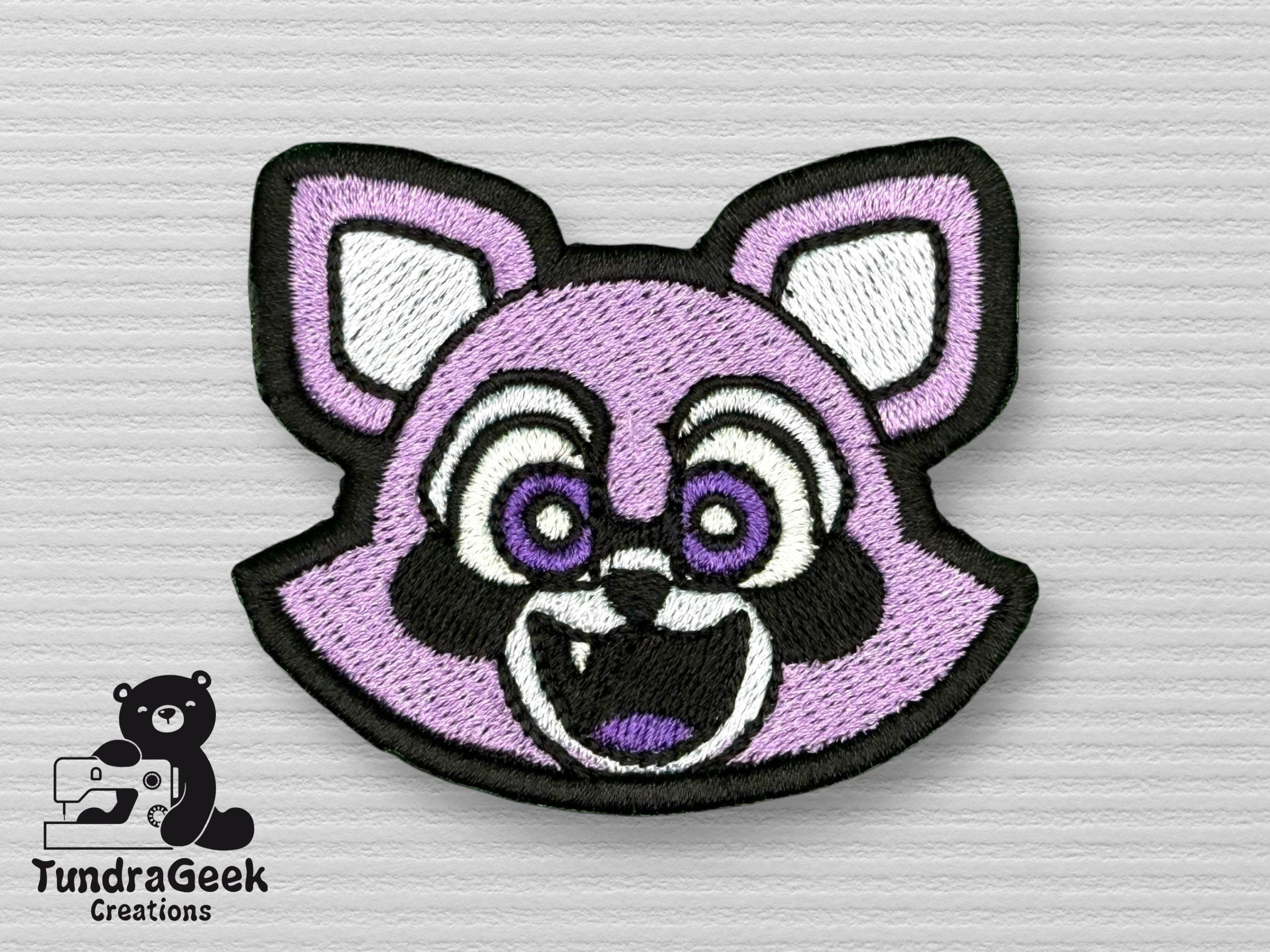 Embroidered patch of a stylized, smiling purple raccoon face with white and black details, displayed on a textured light gray surface with the TundraGeek Creations logo in the corner.
