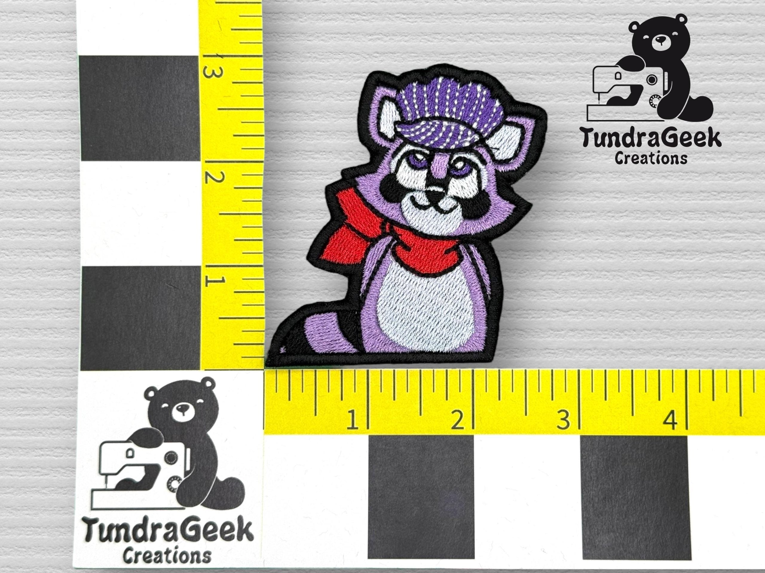 An embroidered patch of a purple raccoon wearing a red scarf and purple hat, measured on a yellow and black ruler grid with the TundraGeek Creations logo visible.
