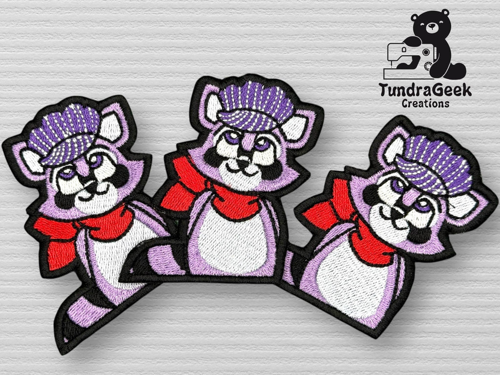 Three identical embroidered patches of a purple raccoon wearing a red scarf and a purple knit hat, displayed on a textured surface next to the TundraGeek Creations logo.