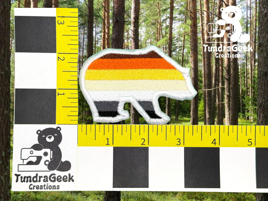 A bear-shaped patch with horizontal stripes in black, gray, white, orange, and yellow is shown next to a ruler for scale on a forest background.