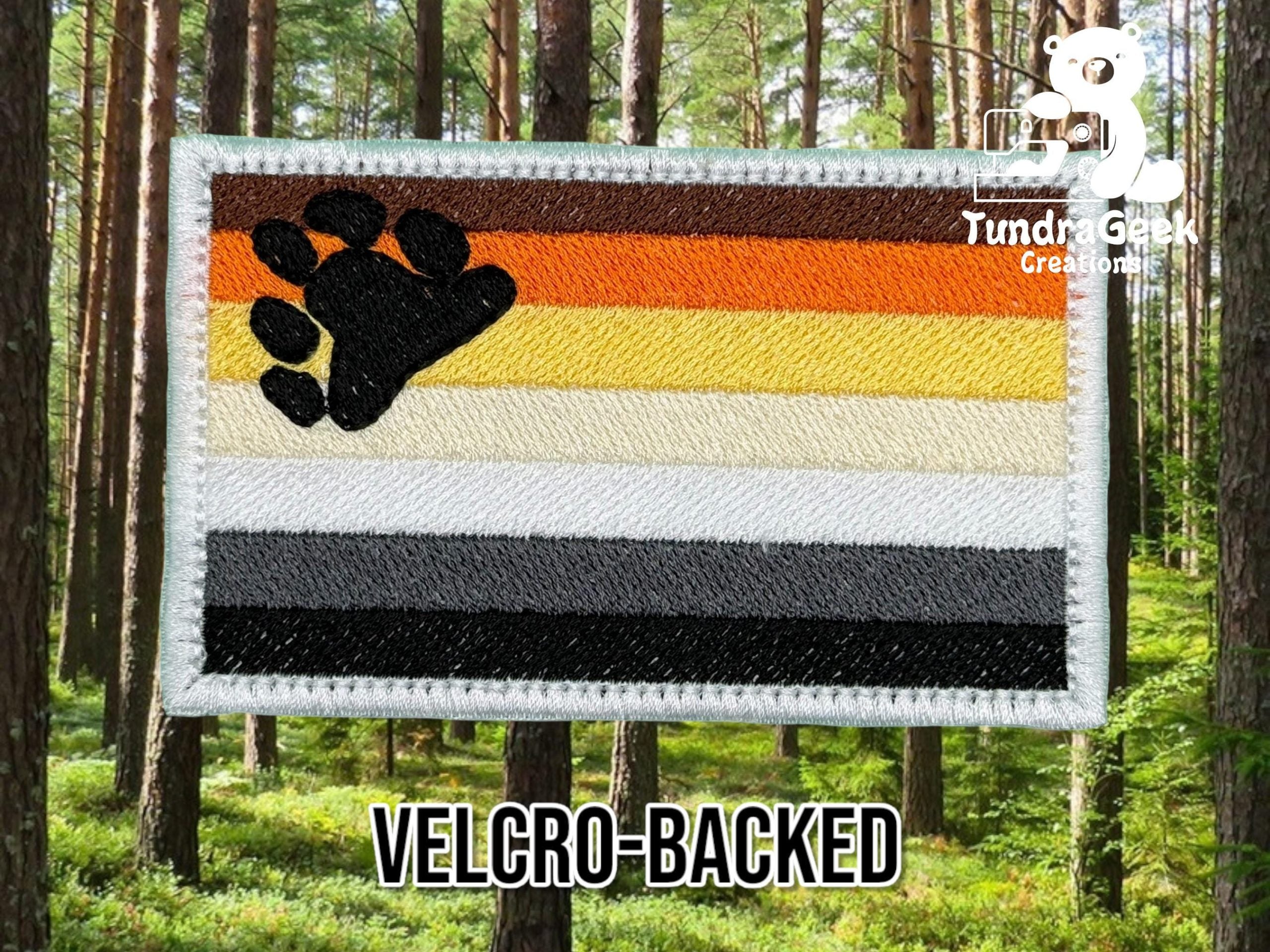 A rectangular embroidered patch with horizontal stripes in brown, orange, yellow, white, gray, and black, featuring a black paw print in the upper left corner. Text reads “VELCRO-BACKED.”.
