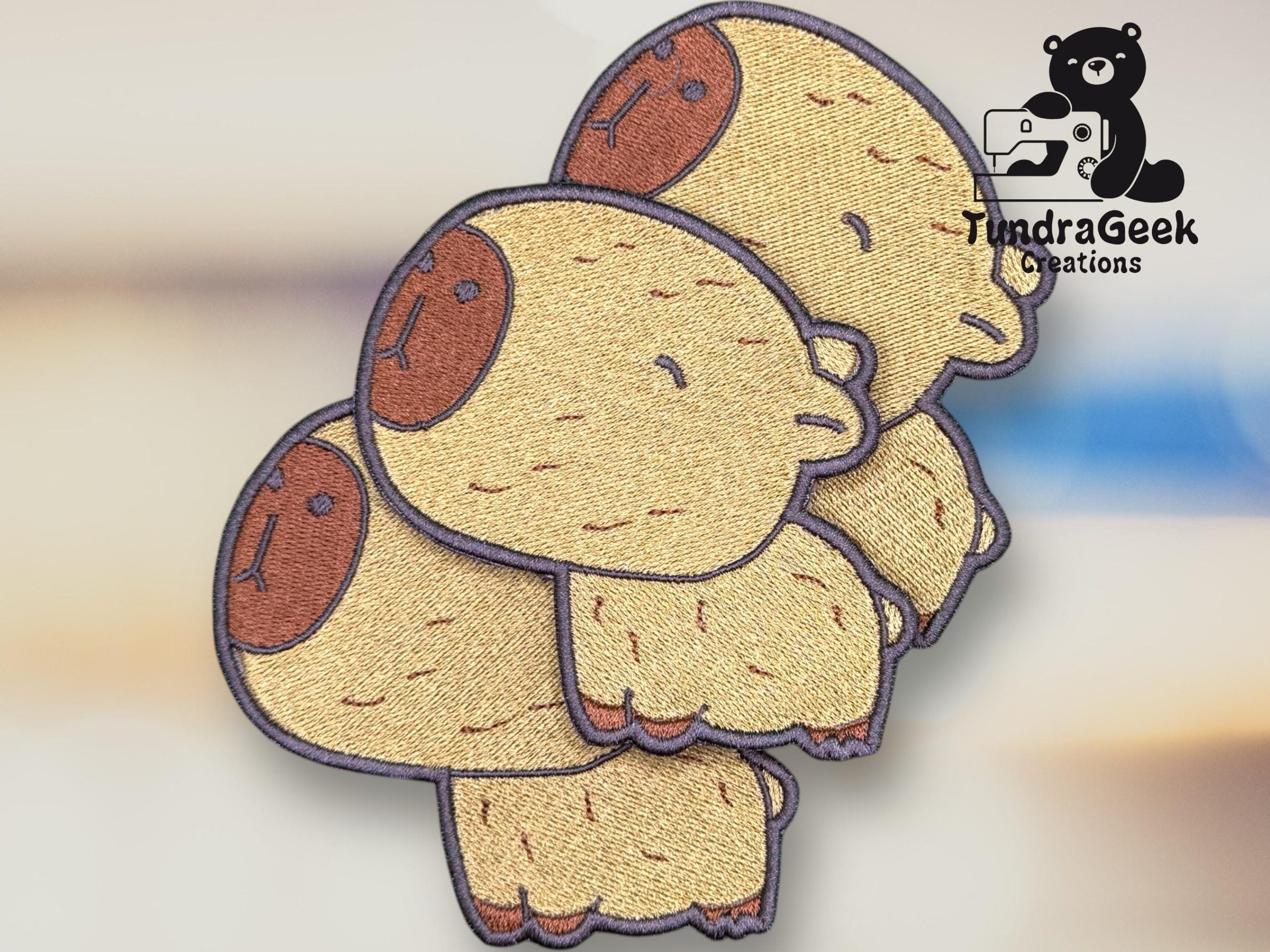 Three embroidered patches feature cartoon capybaras stacked on top of each other. The TundraGeek Creations logo appears in the upper right corner.