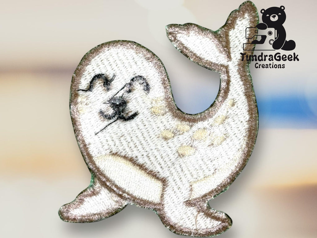 A shiny embroidered patch of a smiling seal with white and gray details, next to the TundraGeek Creations logo featuring a bear holding a sewing machine.