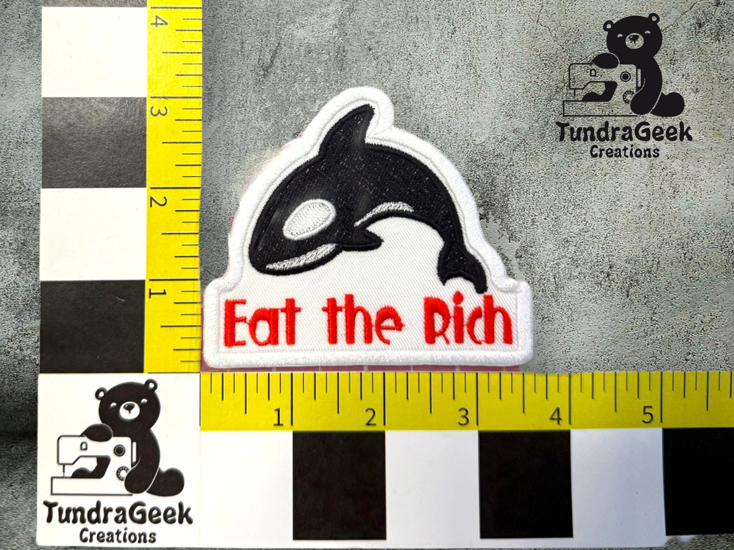 A patch featuring a black and white orca with the text Eat the Rich in red letters, surrounded by measurement guides and TundraGeek Creations logos.