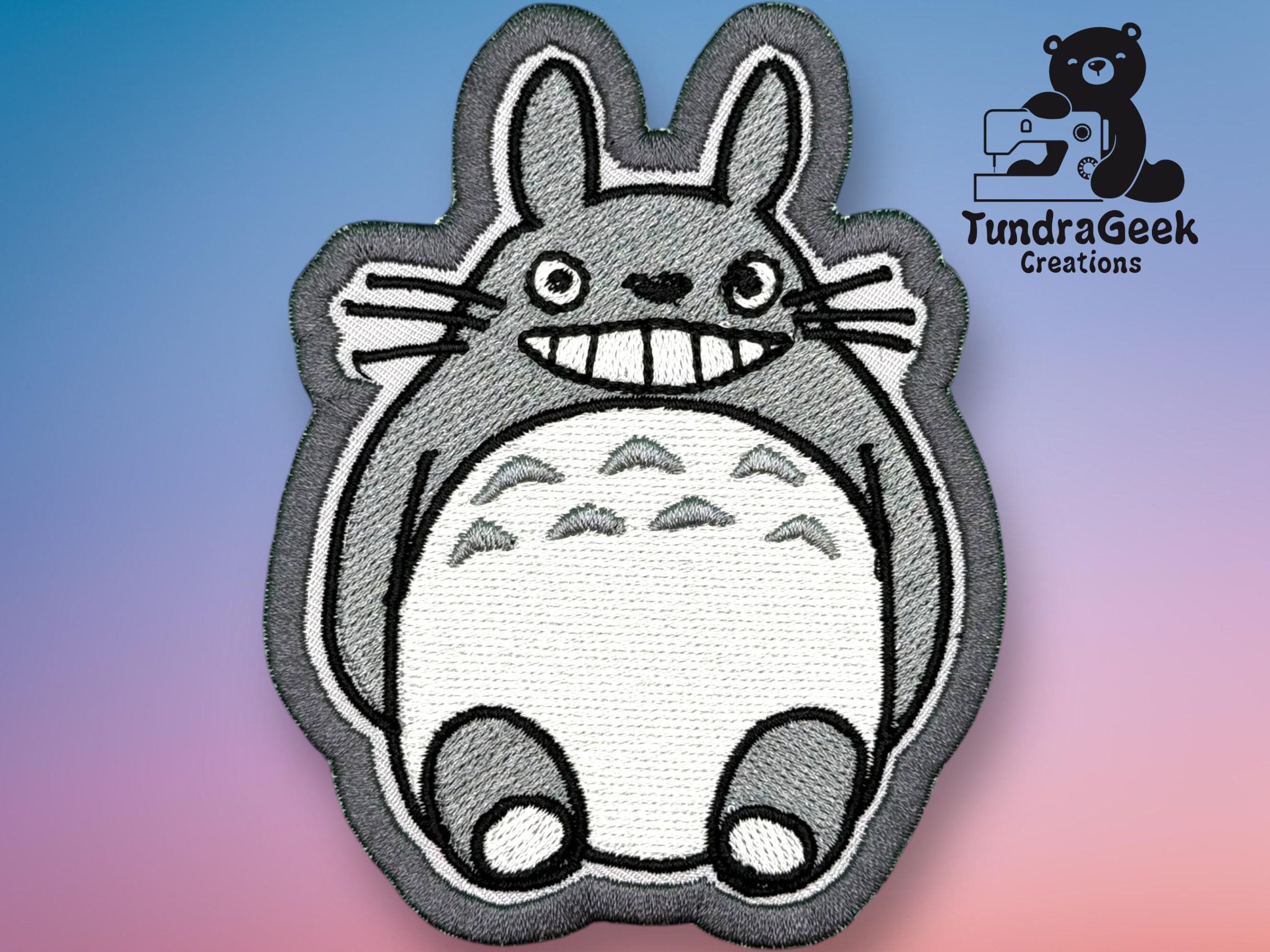 Embroidered patch featuring a cartoon gray creature with large eyes and a wide smile, displayed on a gradient background with TundraGeek Creations logo in the upper right corner.