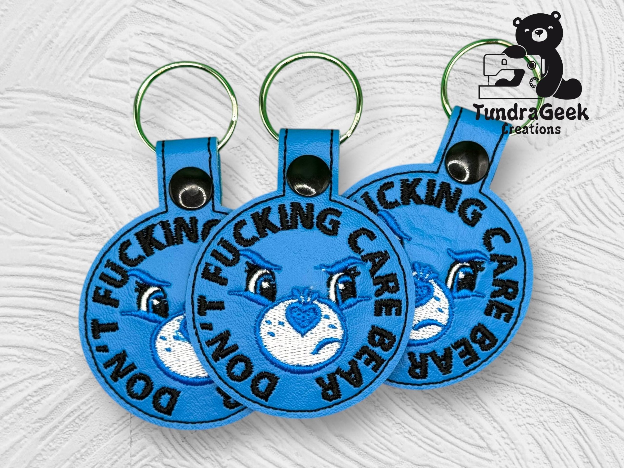 Three blue embroidered keychains display an angry cartoon bear and the words DONT F***ING CARE BEAR. The Tundra Geek Creations logo is in the upper right corner.