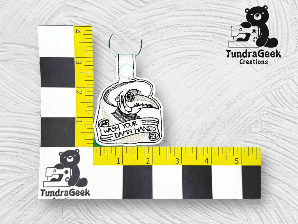 A keychain with an embroidery of hands being washed and the text WASH YOUR DAMN HANDS, displayed next to a ruler for scale, with the TundraGeek Creations logo in the corner.