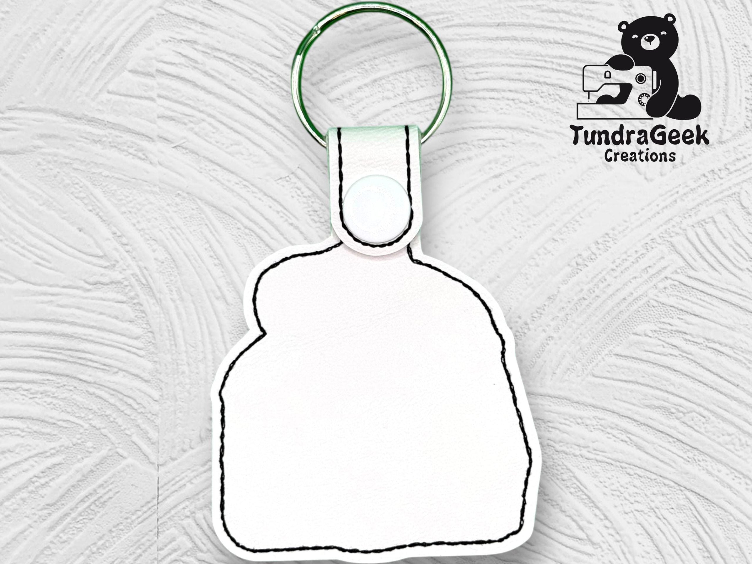 A blank white keychain with black stitching, featuring a metal ring, is displayed on a textured white background. The TundraGeek Creations logo appears in the top right corner.
