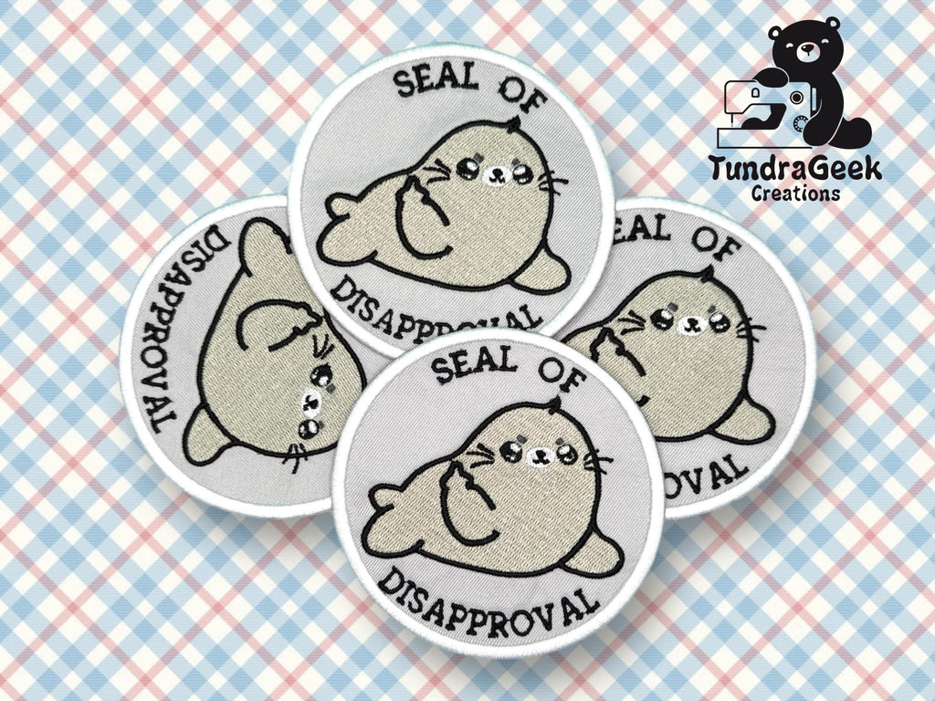 Four embroidered patches featuring a cartoon seal with the text Seal of Disapproval are arranged in a fan shape on a plaid background. TundraGeek Creations logo is in the corner.