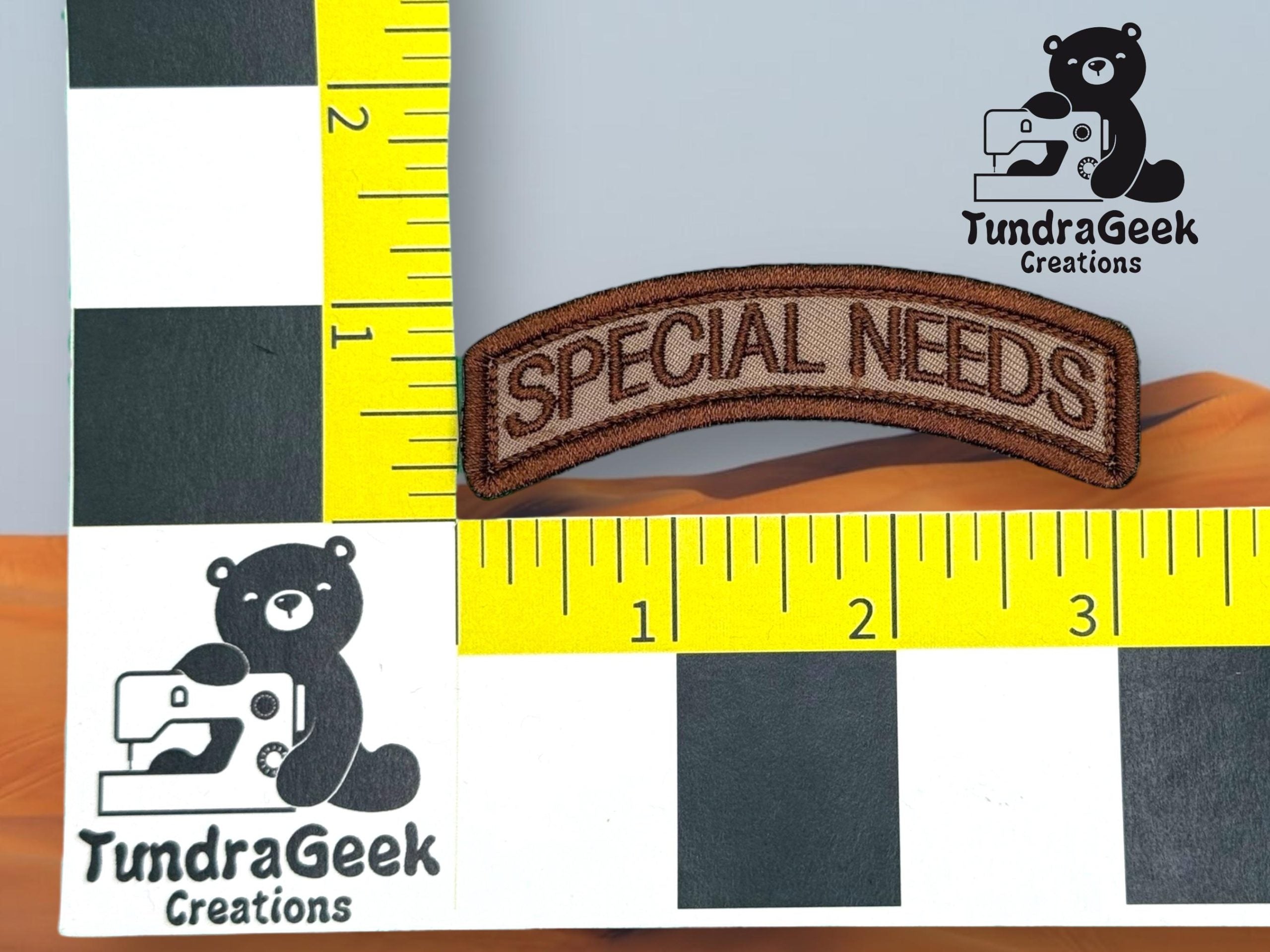 A brown embroidered patch reading SPECIAL NEEDS is placed on a measurement grid with TundraGeek Creations logos featuring a bear with a sewing machine.