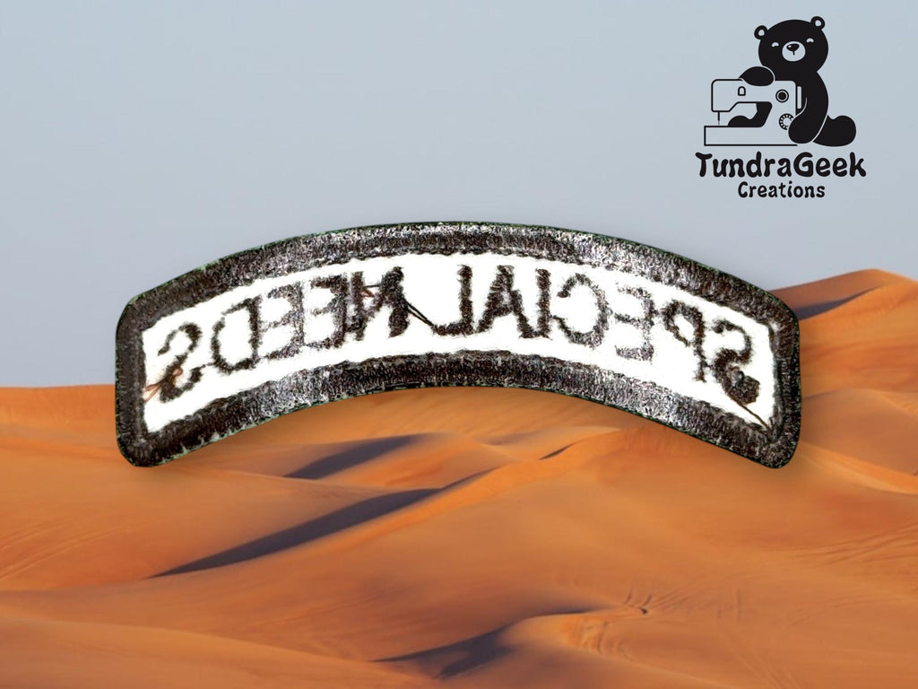 A curved fabric patch with the mirrored text SPECIAL NEEDS lies over a desert sand dune background, with the TundraGeek Creations logo in the top right corner.