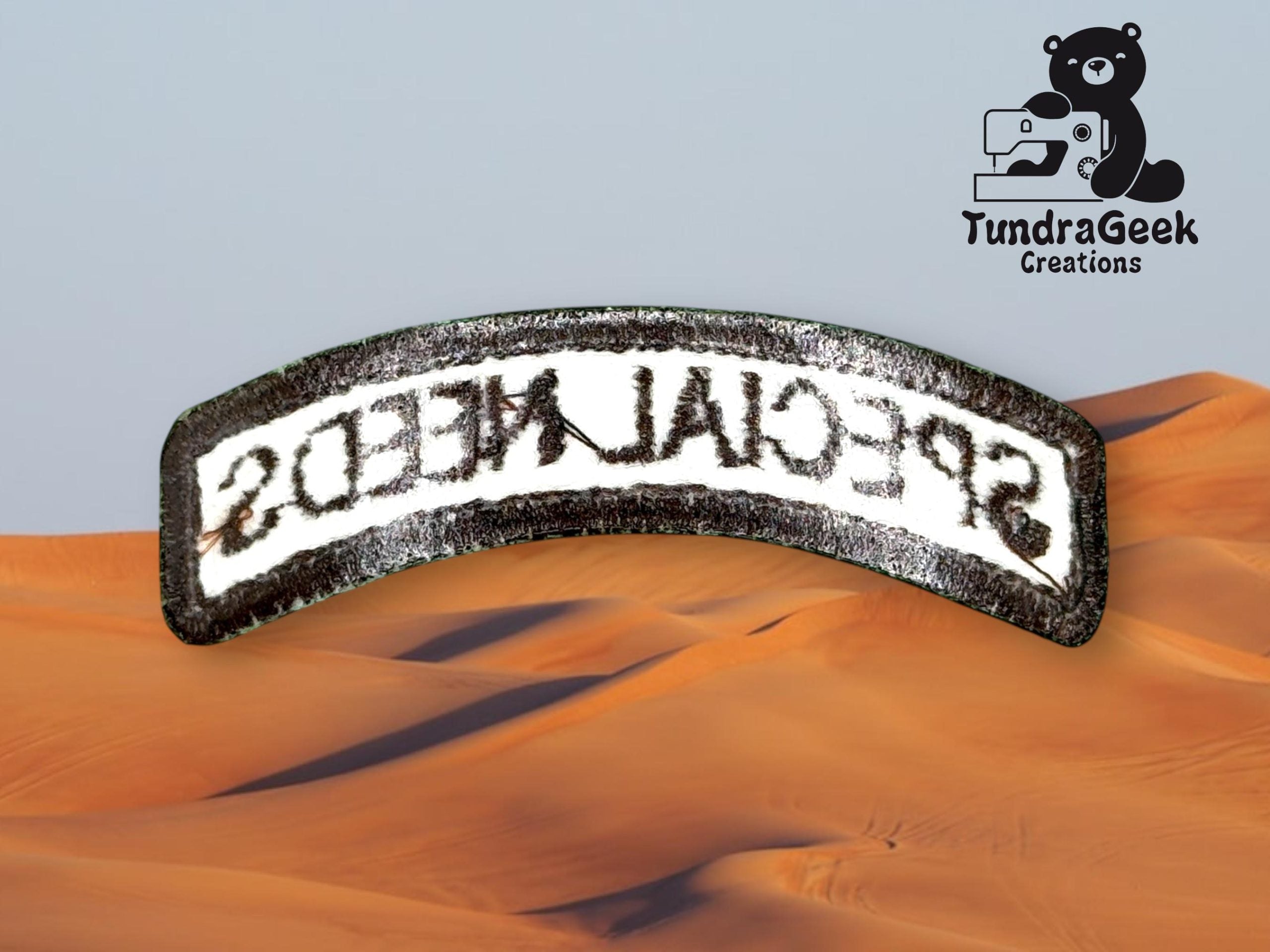 A curved fabric patch with the mirrored text SPECIAL NEEDS lies over a desert sand dune background, with the TundraGeek Creations logo in the top right corner.