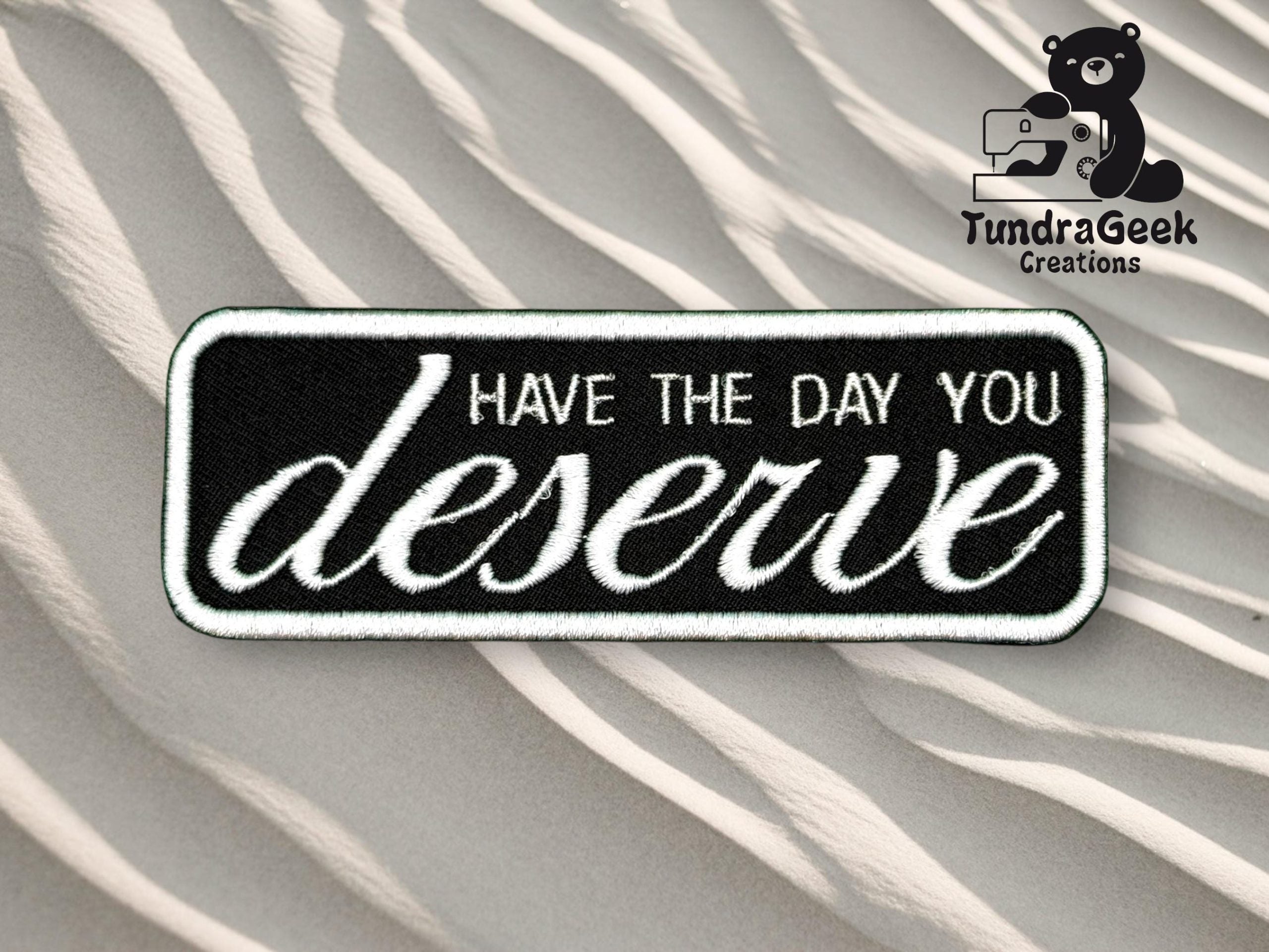 A rectangular black and white patch reads Have the day you deserve on a sand dune background with the TundraGeek Creations logo in the top right corner.