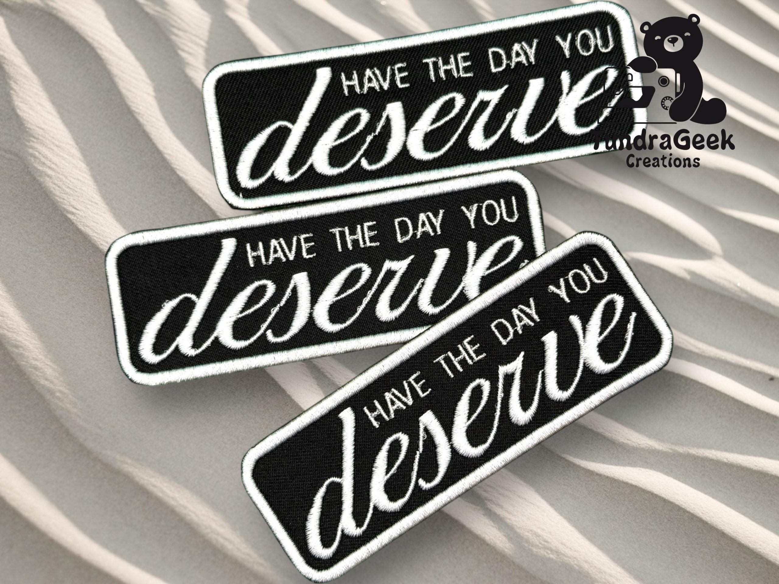 Three black and white patches with the phrase Have the day you deserve are displayed on a wavy, sand-like background. The Tundra Geek Creations logo is in the top right corner.