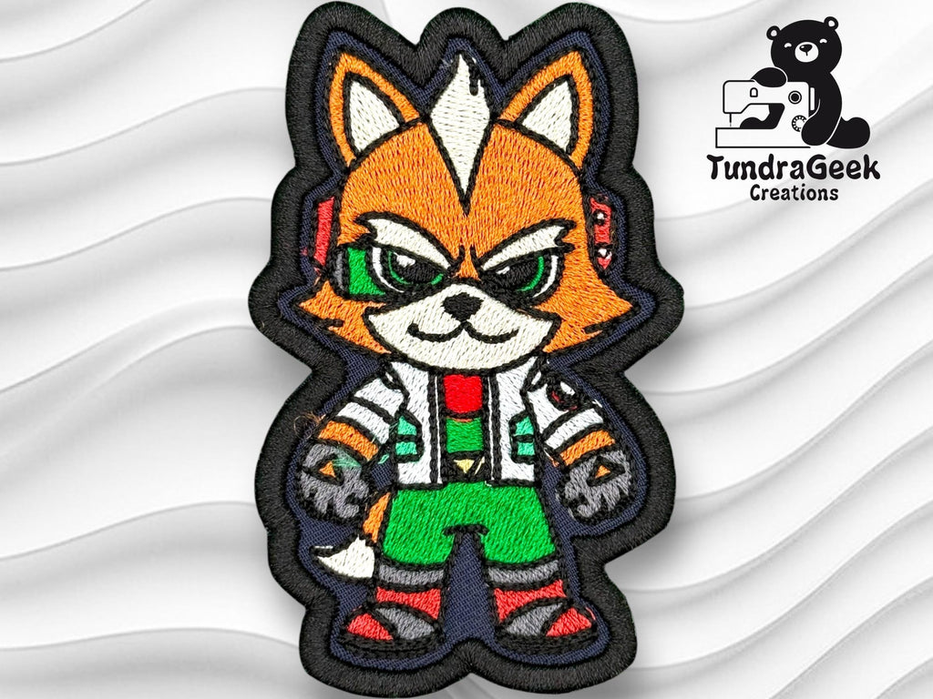 Embroidered patch featuring a cartoon fox with orange fur, green pants, and tech accessories, on a wavy white background. TundraGeek Creations logo appears in the top right corner.
