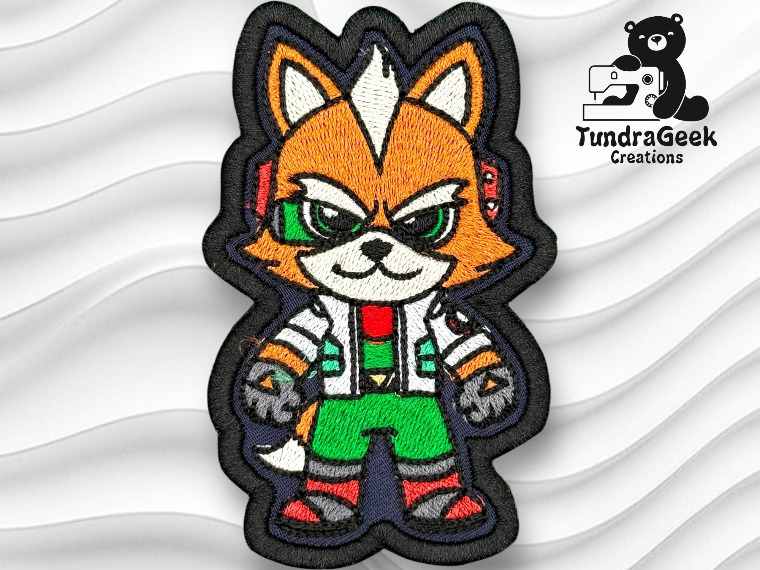 Embroidered patch featuring a cartoon fox with orange fur, green pants, and tech accessories, on a wavy white background. TundraGeek Creations logo appears in the top right corner.