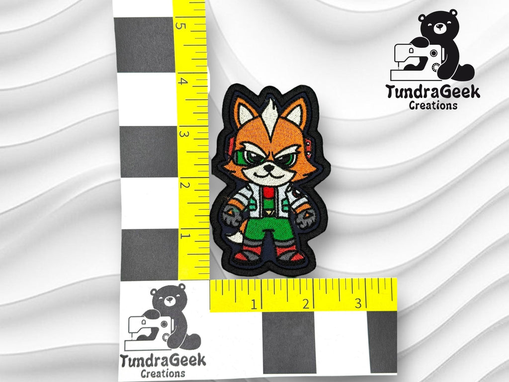 Embroidered patch of an anthropomorphic fox in green overalls on a white background, next to a ruler for scale, labeled with TundraGeek Creations logo.