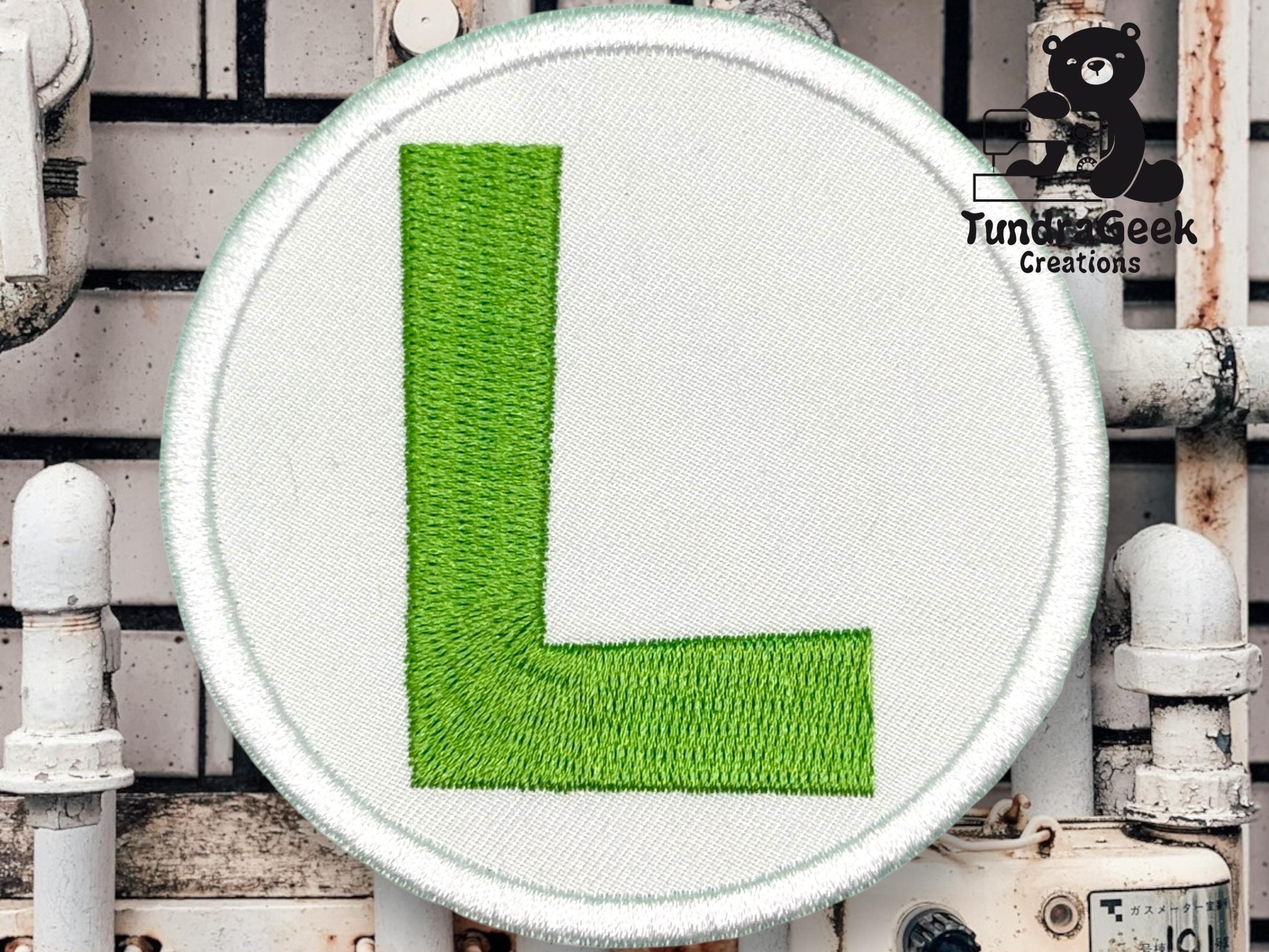 A circular fabric patch with a large green letter L on a white background, with the Tundra Geek Creations logo in the top right corner.