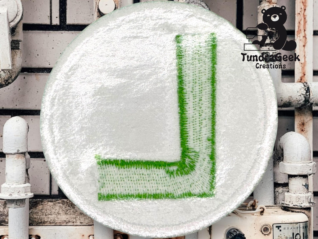 A round white patch featuring a green embroidered letter L rests against a background of white metal pipes. A cartoon bear logo reading Tunda Geek Creations is in the upper right corner.