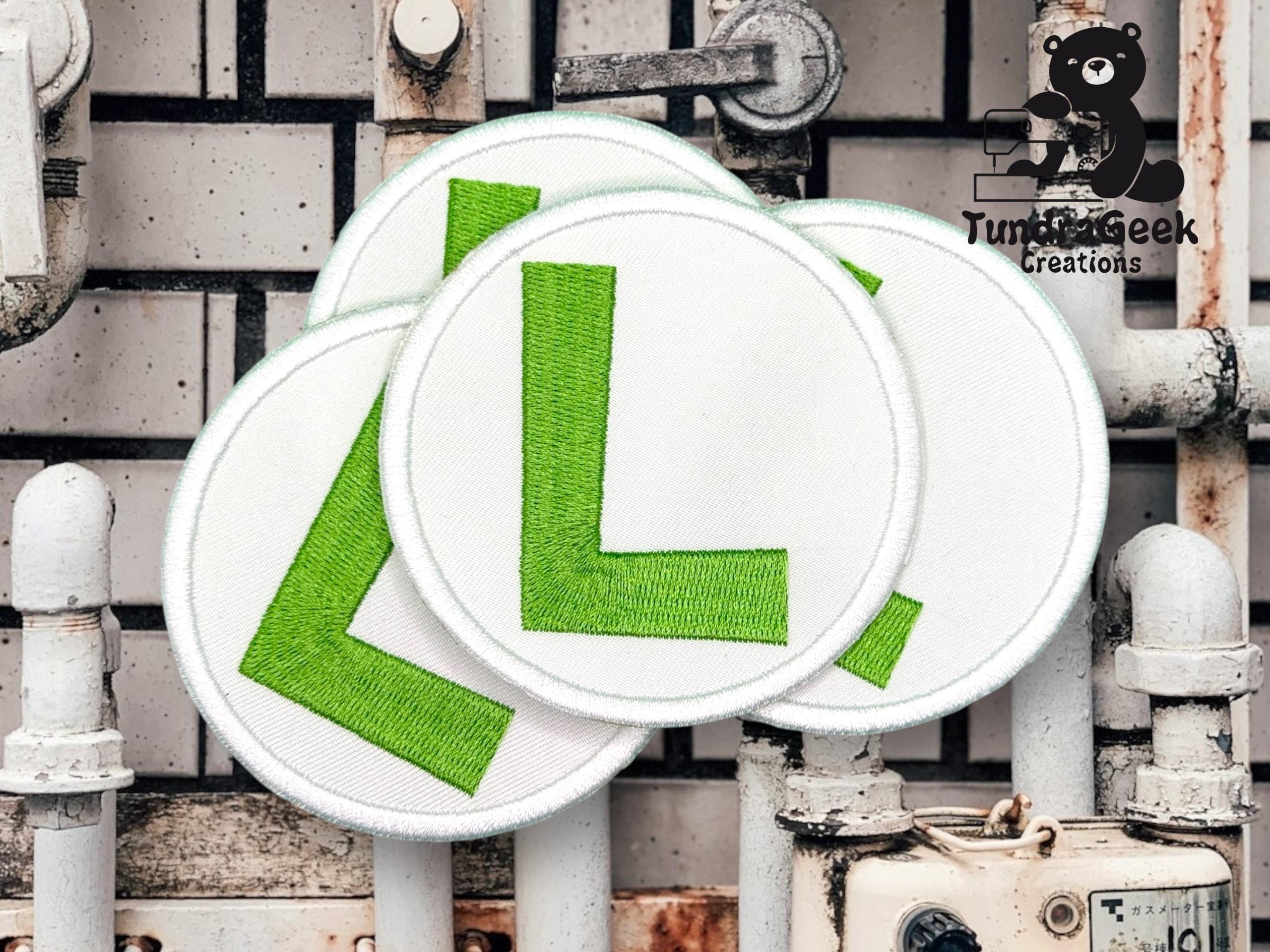 Four circular patches with large green L letters are arranged overlapping on a white industrial pipe background. A cartoon bear and TurtleGeek Creations logo are visible in the top right corner.