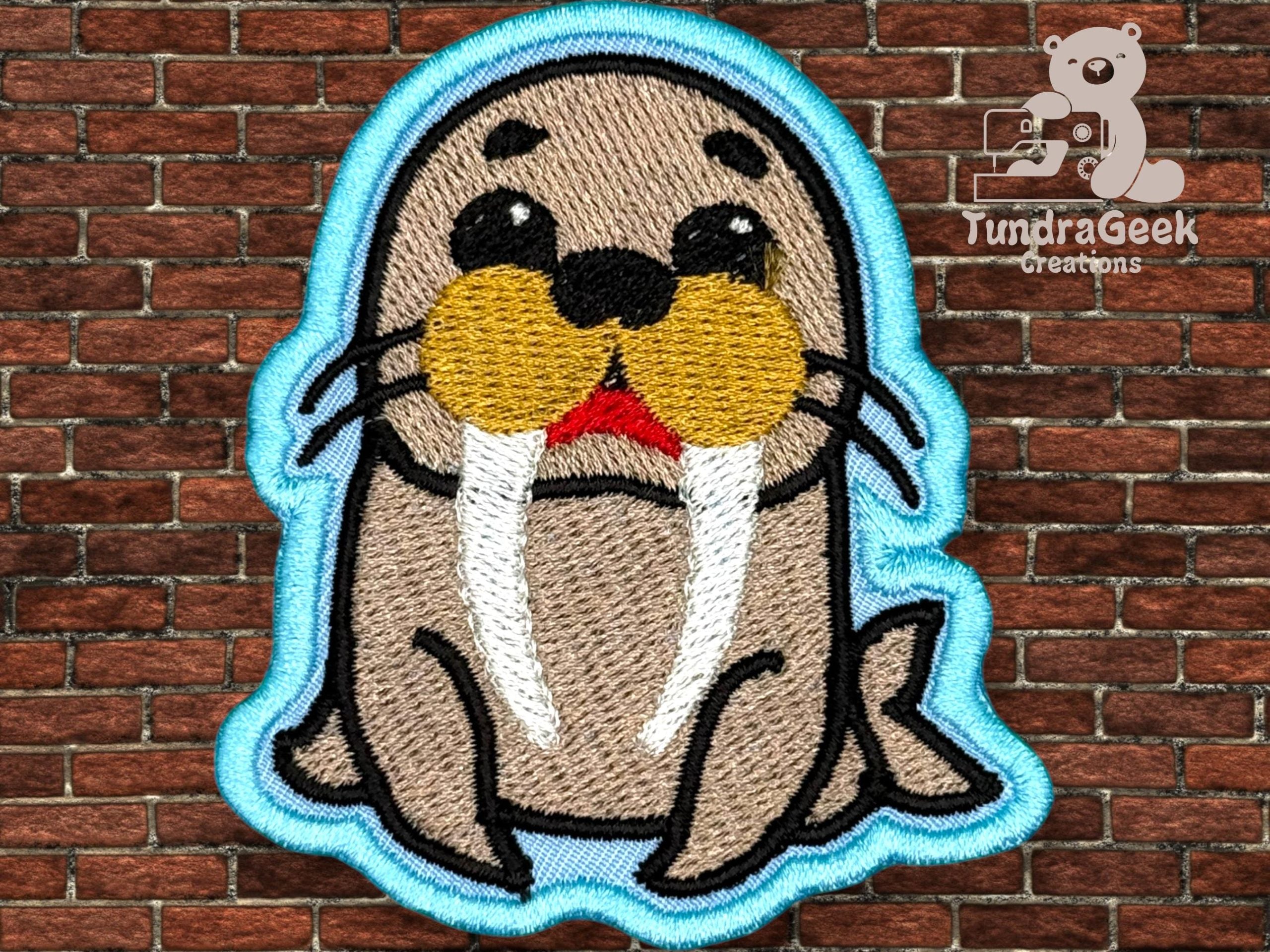 Embroidered patch of a cartoon walrus with prominent tusks and yellow whiskers, outlined in blue, displayed on a brick wall background. Logo reads TundraGeek Creations with a bear graphic.