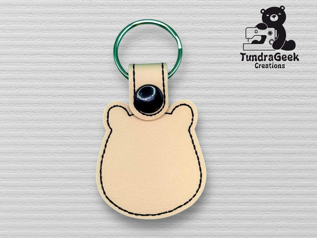 A bear-shaped beige leather keychain with black stitching and a green metal ring, displayed on a textured background. TundraGeek Creations logo is visible in the upper right corner.