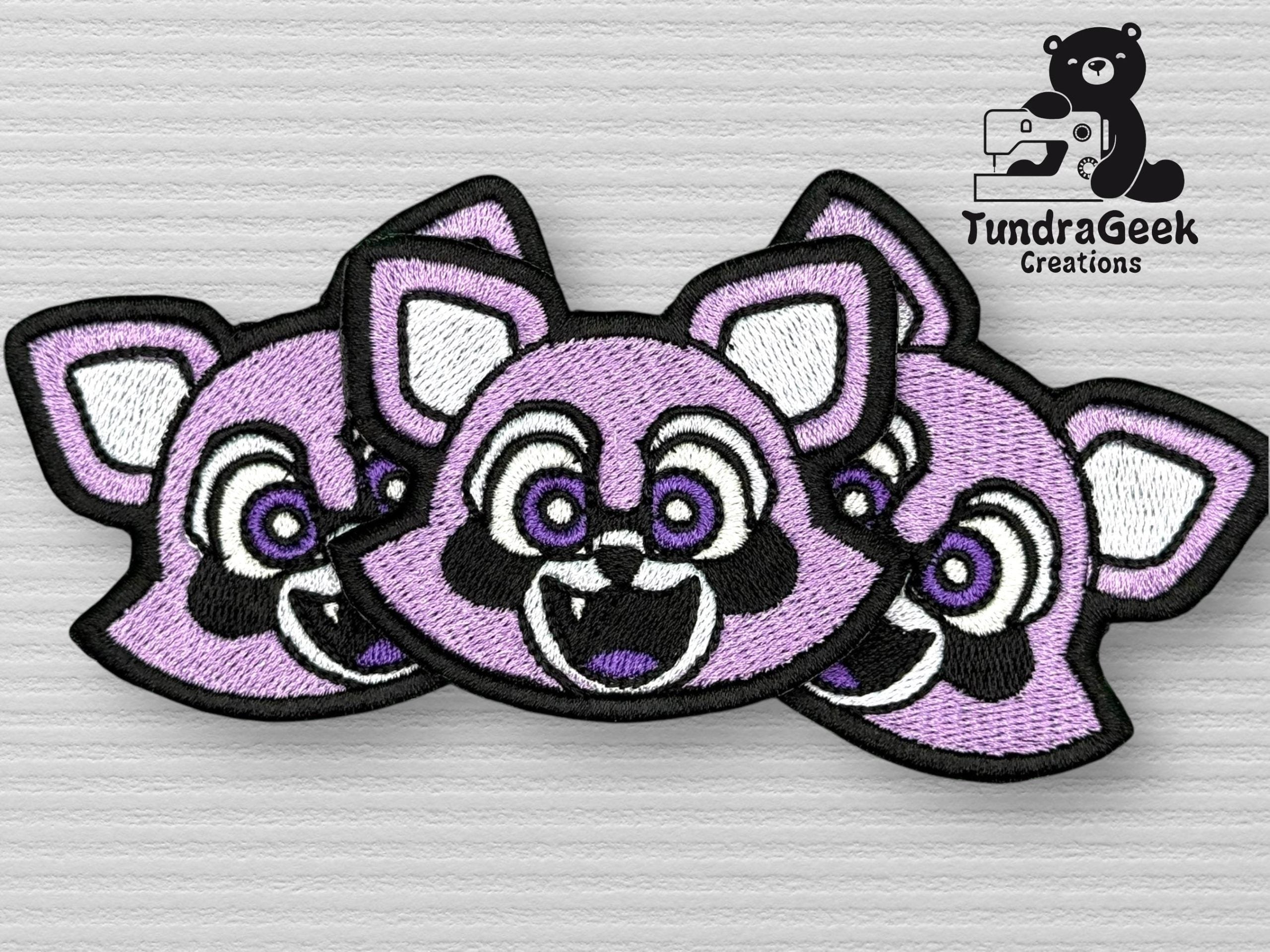 Three purple raccoon face embroidered patches overlap on a gray textured surface. The TundraGeek Creations logo with a bear and text appears in the top right corner.