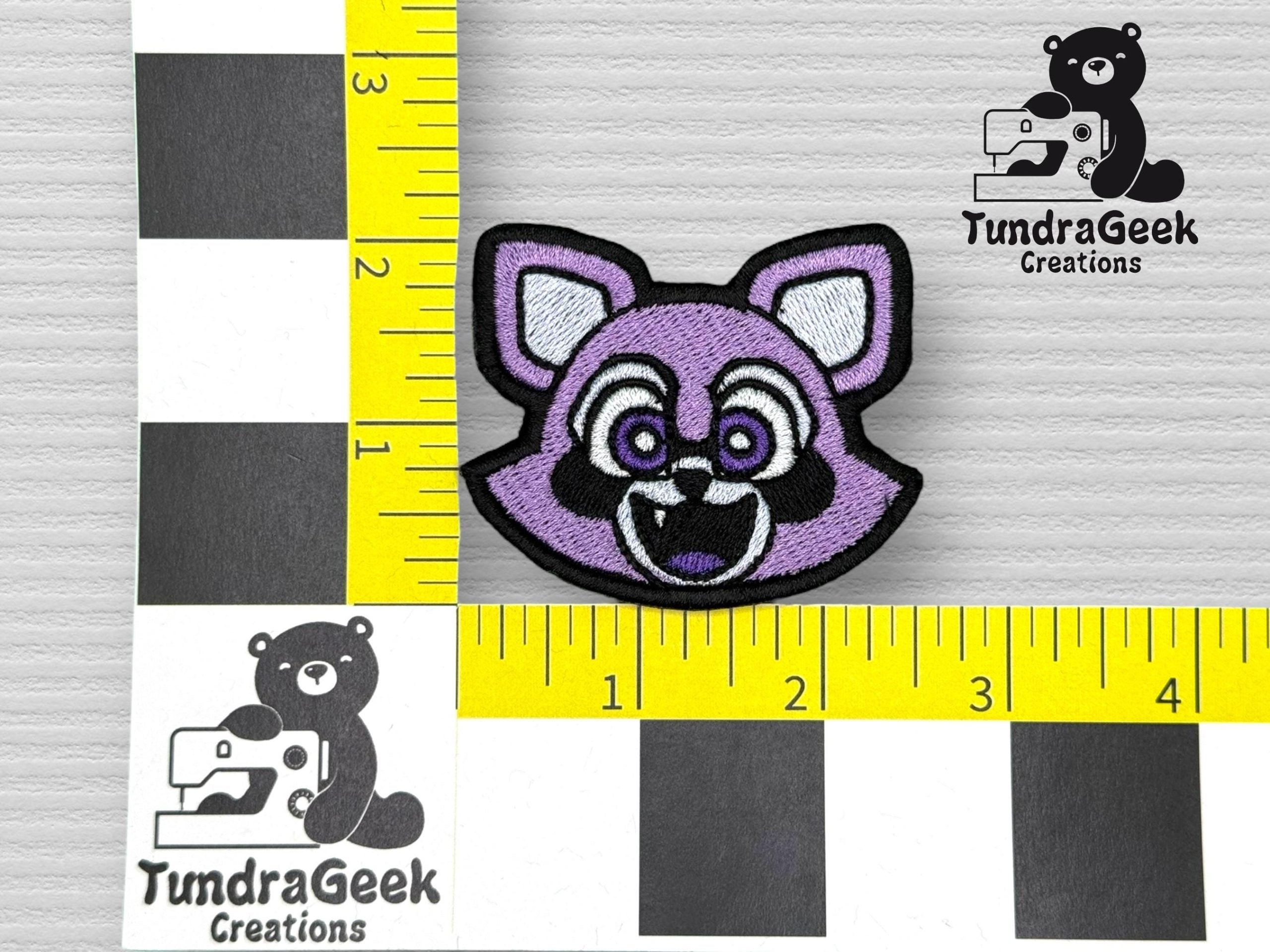 Embroidered patch of a stylized purple raccoon head placed on a ruler grid for size reference, next to the TundraGeek Creations logo.