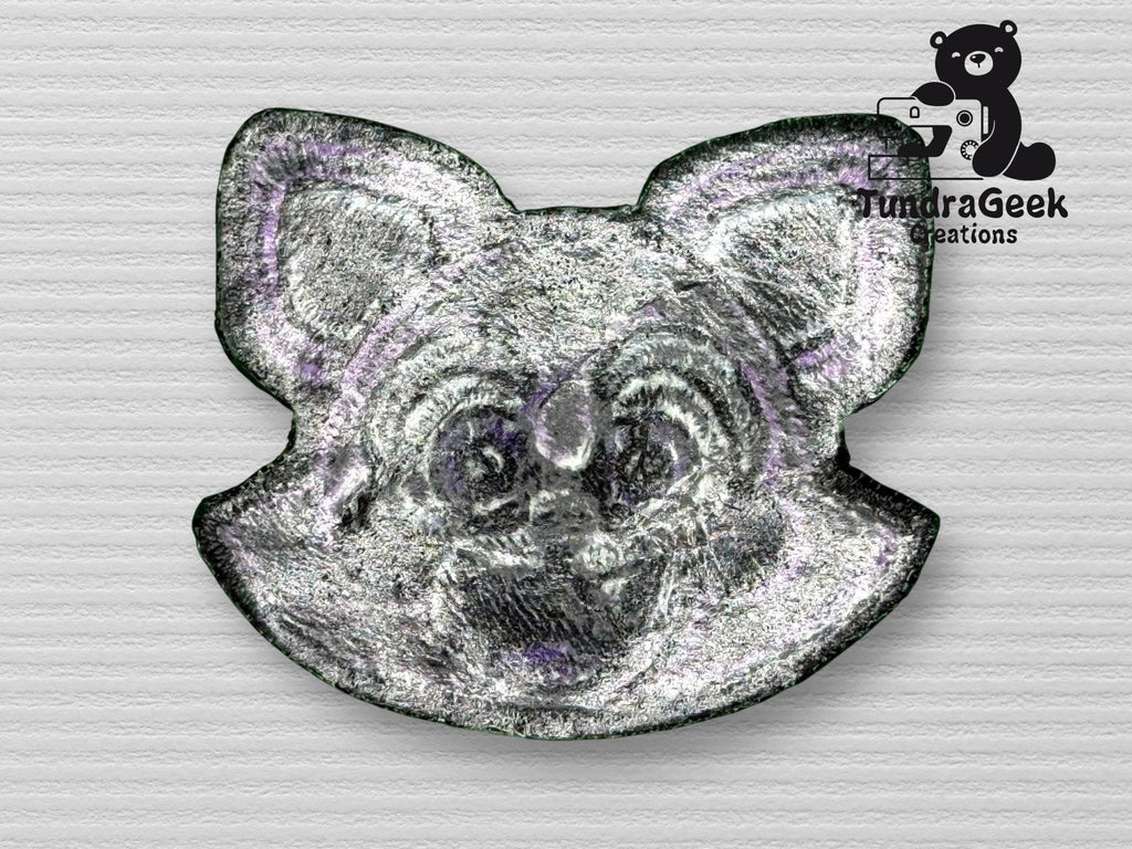 A metallic pin shaped like a cartoon animals face, possibly a cat or fox, with the TundraGeek Creations logo featuring a bear holding a game controller in the top right corner.
