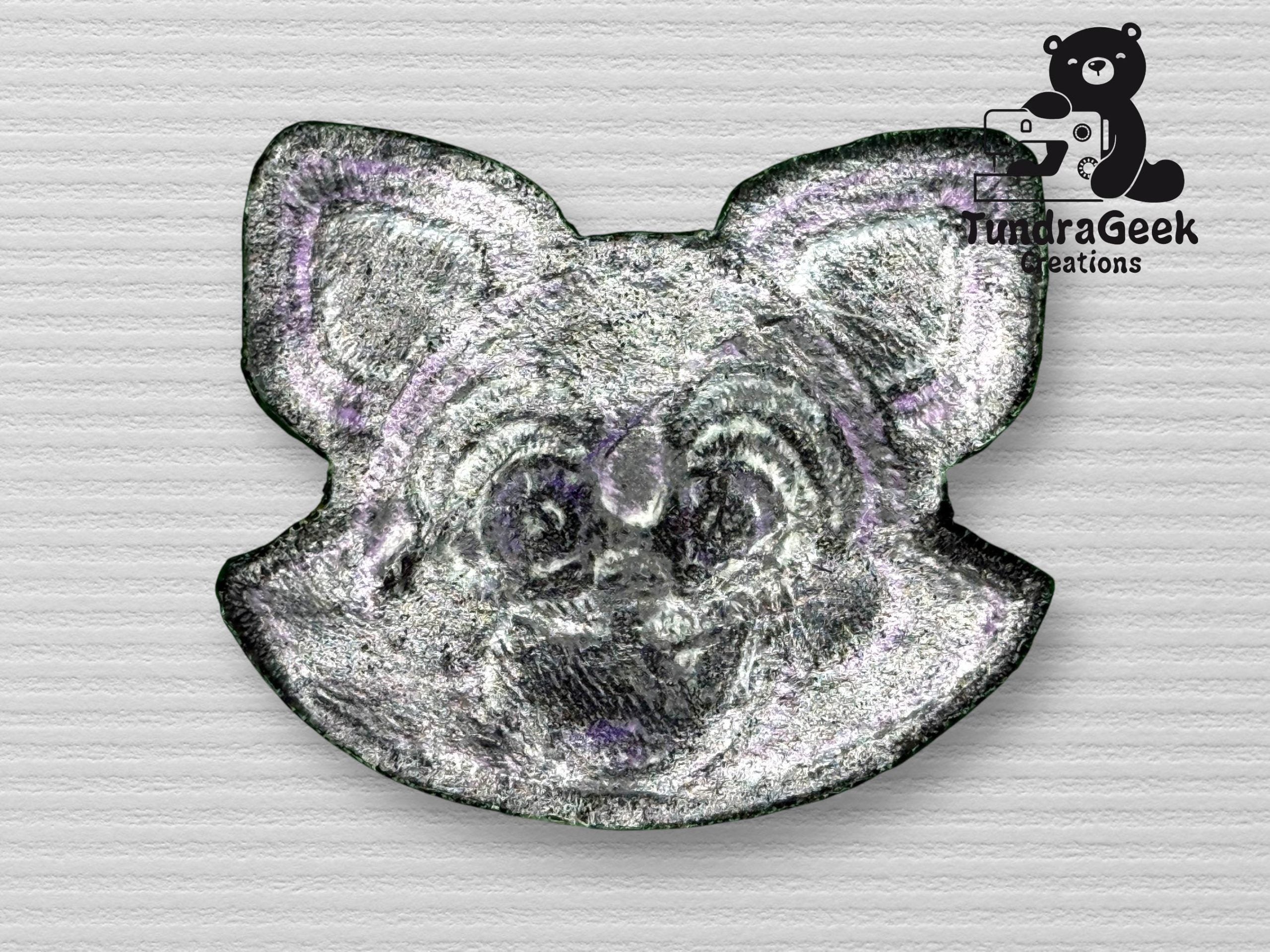 A metallic pin shaped like a cartoon animals face, possibly a cat or fox, with the TundraGeek Creations logo featuring a bear holding a game controller in the top right corner.