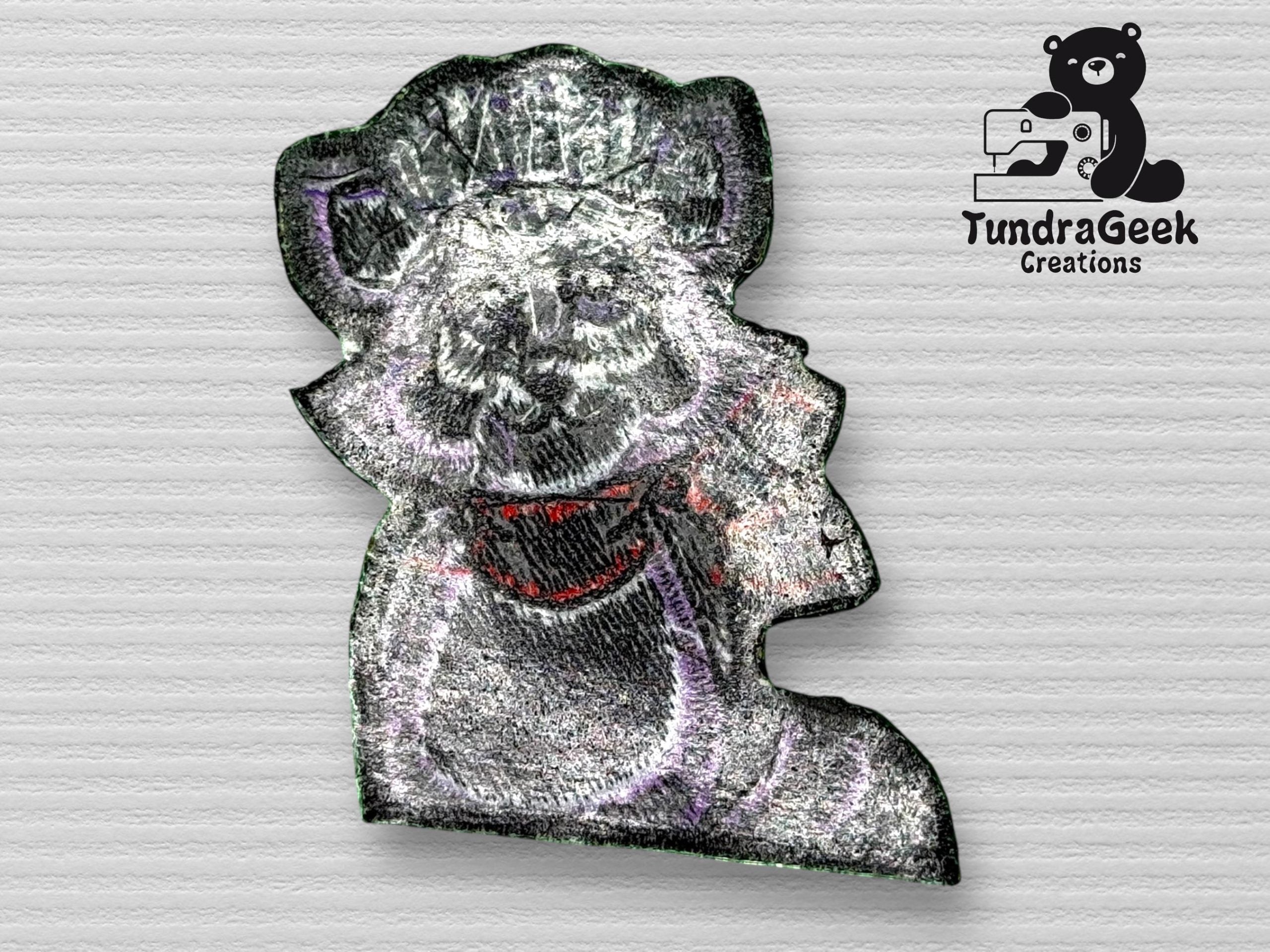 Embroidered patch of a raccoon wearing a chef hat and red scarf, with TundraGeek Creations logo featuring a bear and sewing machine in the upper right corner.