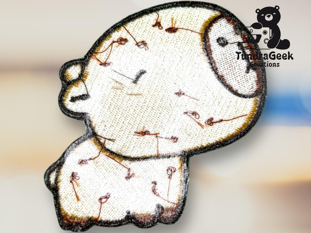Embroidered patch of a stylized, round animal with stitches and seams, shown against a blurred background with a TundraGeek Creations watermark.