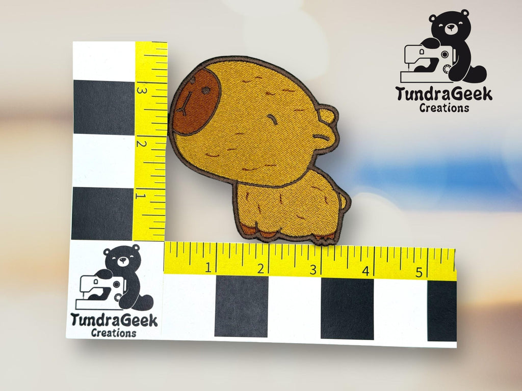 A brown bear patch about 3 inches tall and 3 inches wide, placed on a measurement grid with the TundraGeek Creations logo in the background.