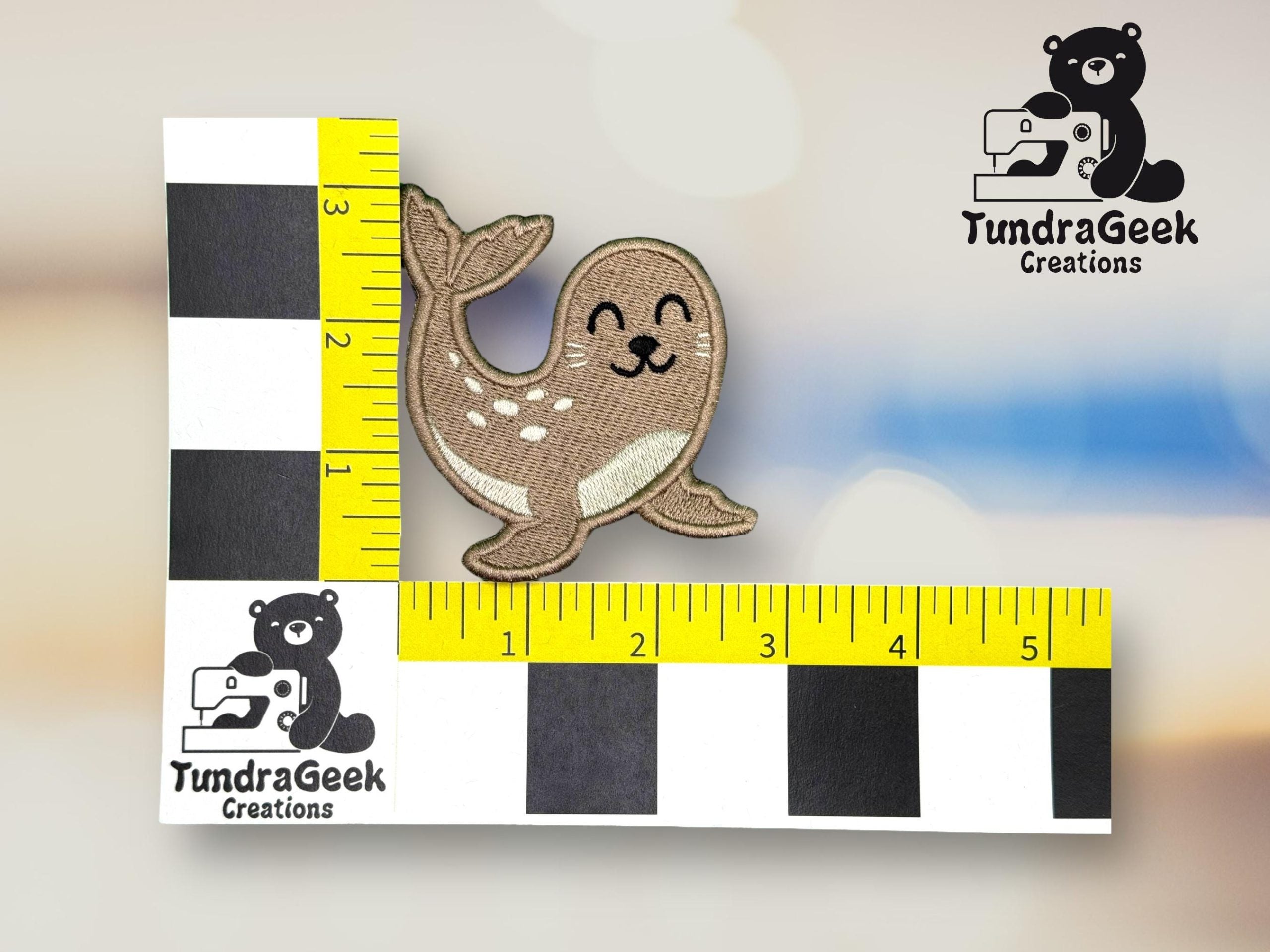 A small embroidered patch of a smiling seal is placed next to a ruler, showing it measures about 2.5 inches wide, with the TundraGeek Creations logo in the background.