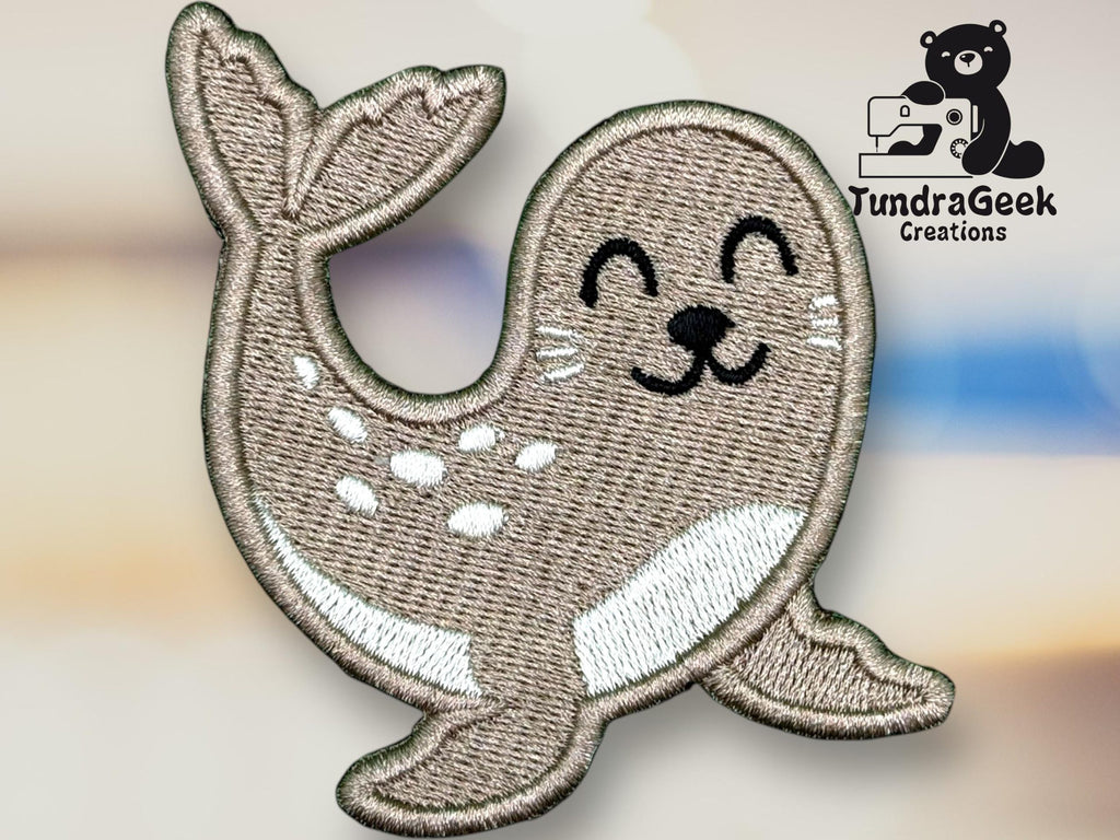 Embroidered patch of a smiling cartoon seal with white spots, placed on a blurred background. Logo for TundraGeek Creations featuring a bear with a sewing machine in the top right corner.
