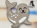 Embroidered patch of a smiling cartoon seal with white spots, placed on a blurred background. Logo for TundraGeek Creations featuring a bear with a sewing machine in the top right corner.