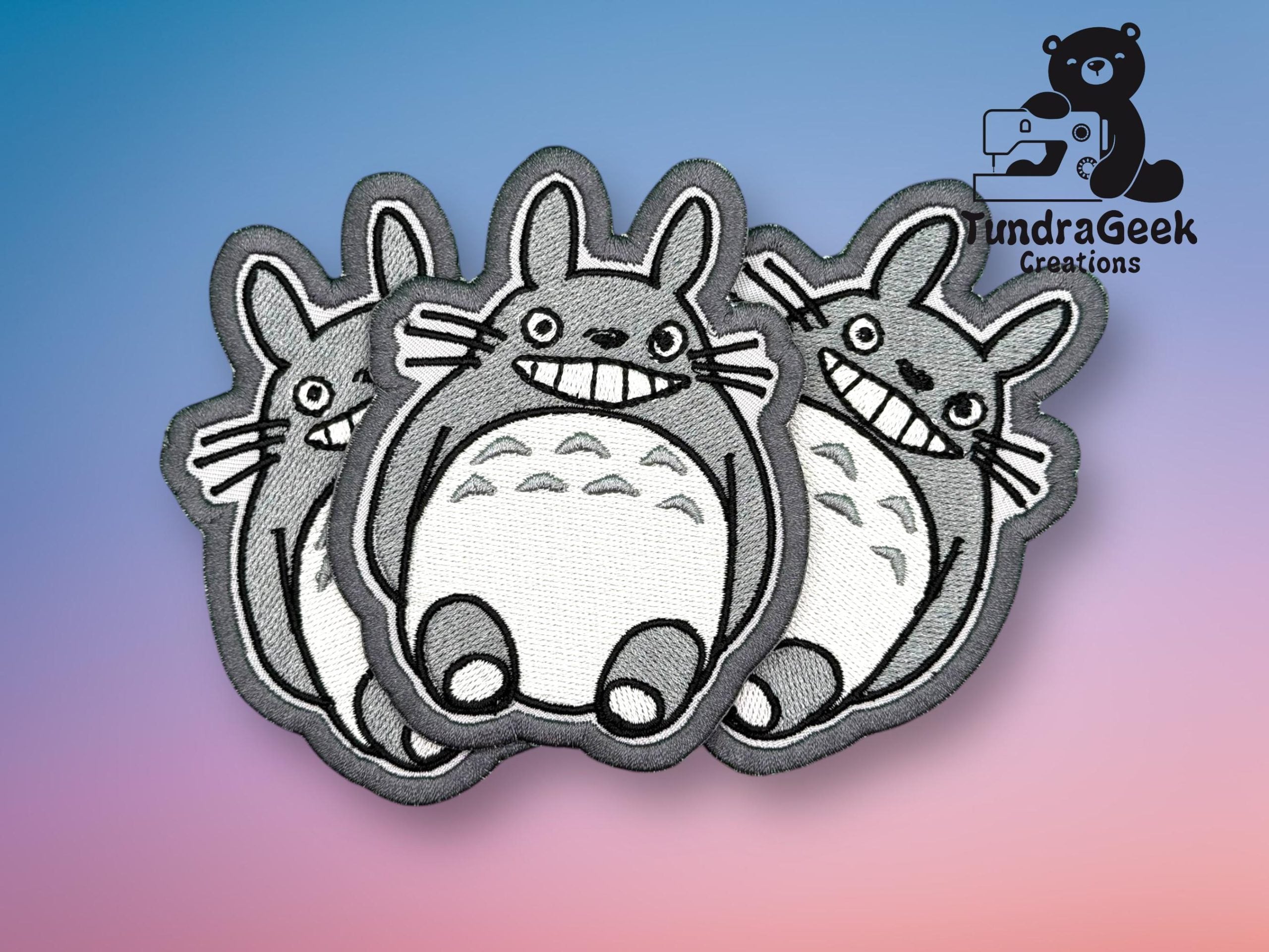 Three embroidered patches featuring a cartoon character with a round body, large grin, and pointy ears, displayed on a gradient blue and pink background with a TundraGeek Creations logo.
