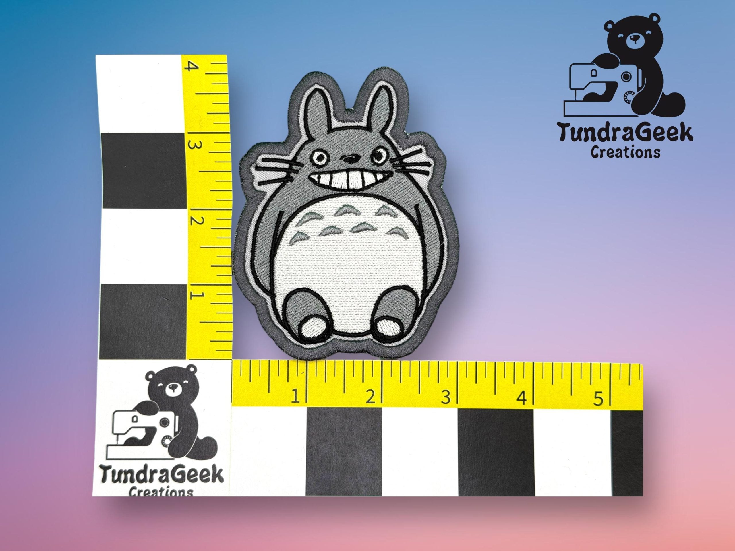 A gray and white Totoro patch is placed next to two rulers for scale, showing the patch is about 3 inches tall. TundraGeek Creations logos are visible on the rulers and background.