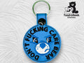 A round blue keychain features an embroidered angry blue bear face and the words DONT FUCKING CARE BEAR around the edge. TundraGeek Creations logo is in the top right corner.