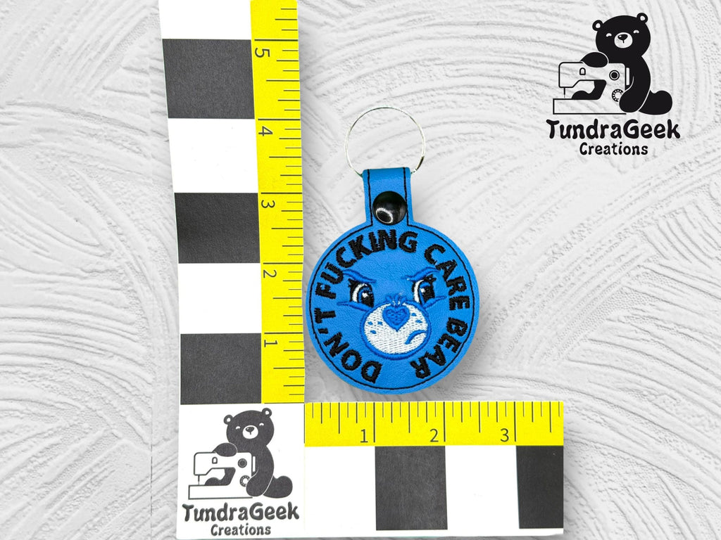A blue keychain with an embroidered bear face and the text DONT FUCKING CARE BEAR, next to rulers showing a size of about 2 inches, with the TundraGeek Creations logo.
