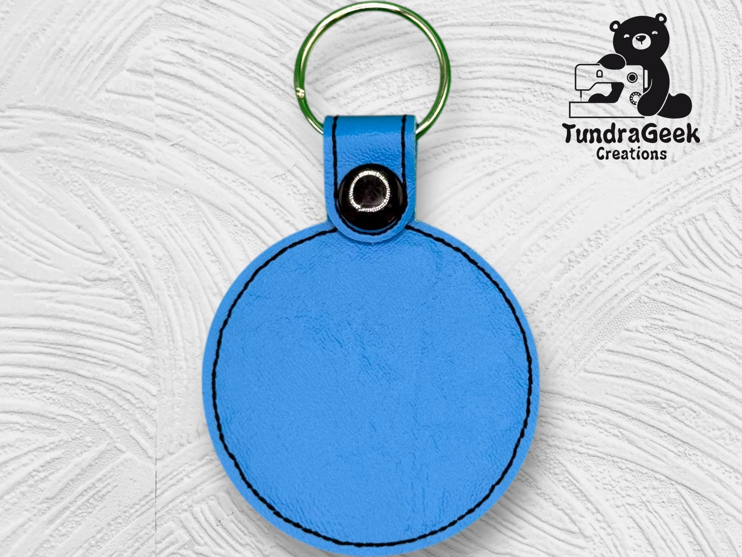 A round blue leather keychain with a green metal ring, displayed on a textured white background, featuring the TundraGeek Creations logo in the top right corner.