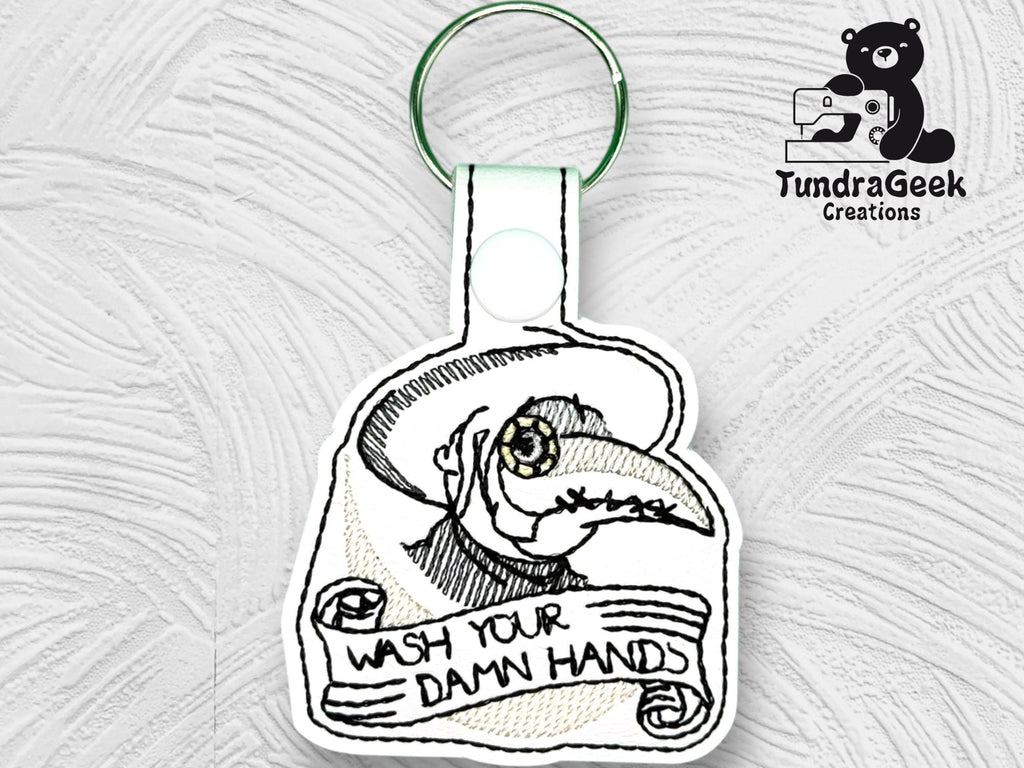 Embroidered keychain featuring a plague doctor illustration and the text Wash Your Damn Hands on a white textured background. TundraGeek Creations logo in the top right corner.