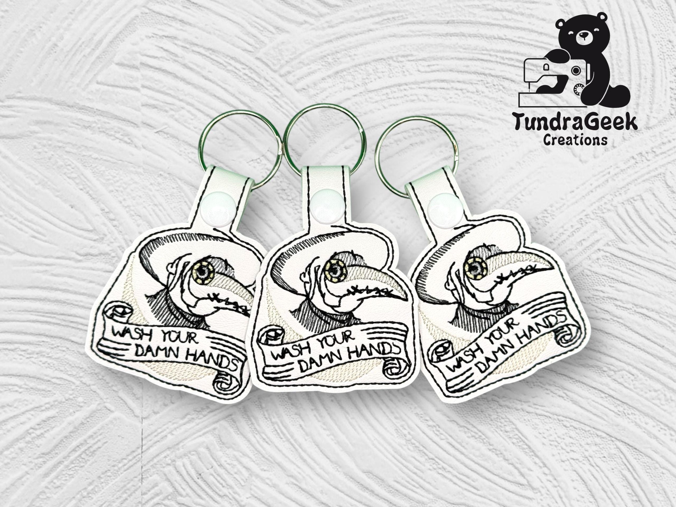 Three identical keychains feature an illustration of a dragon and the text WASH YOUR DAMN HANDS, with the TundraGeek Creations logo in the top right corner.