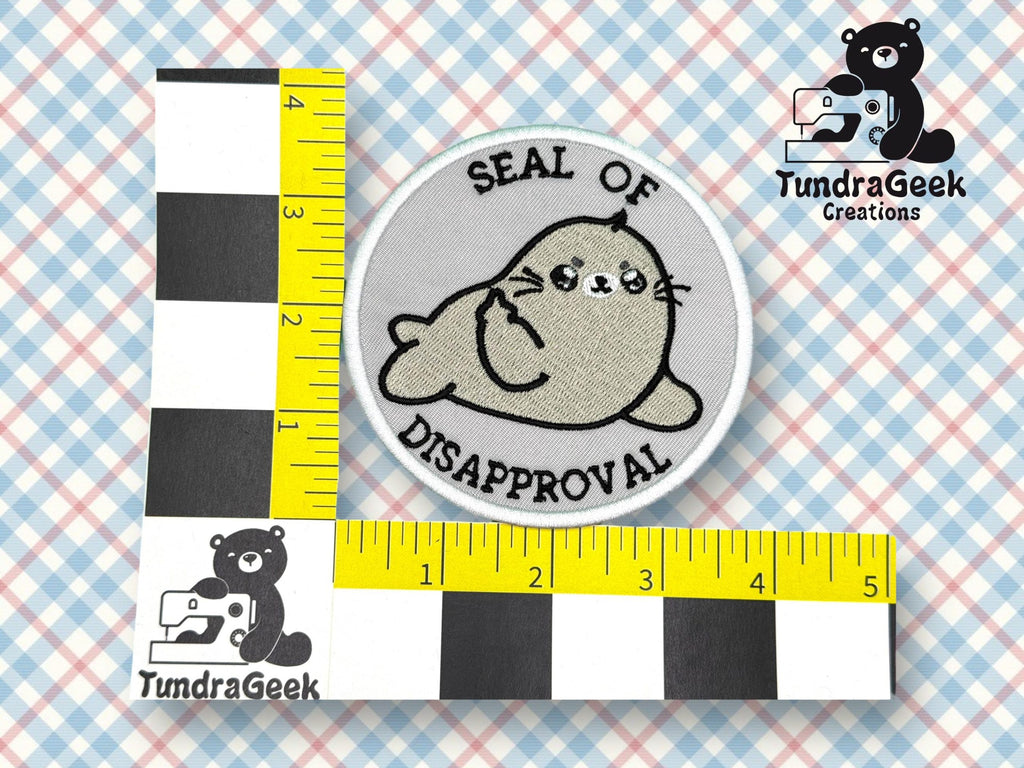 A round embroidered patch with a cartoon seal and the text Seal of Disapproval is placed next to a measuring scale on a checkered background.