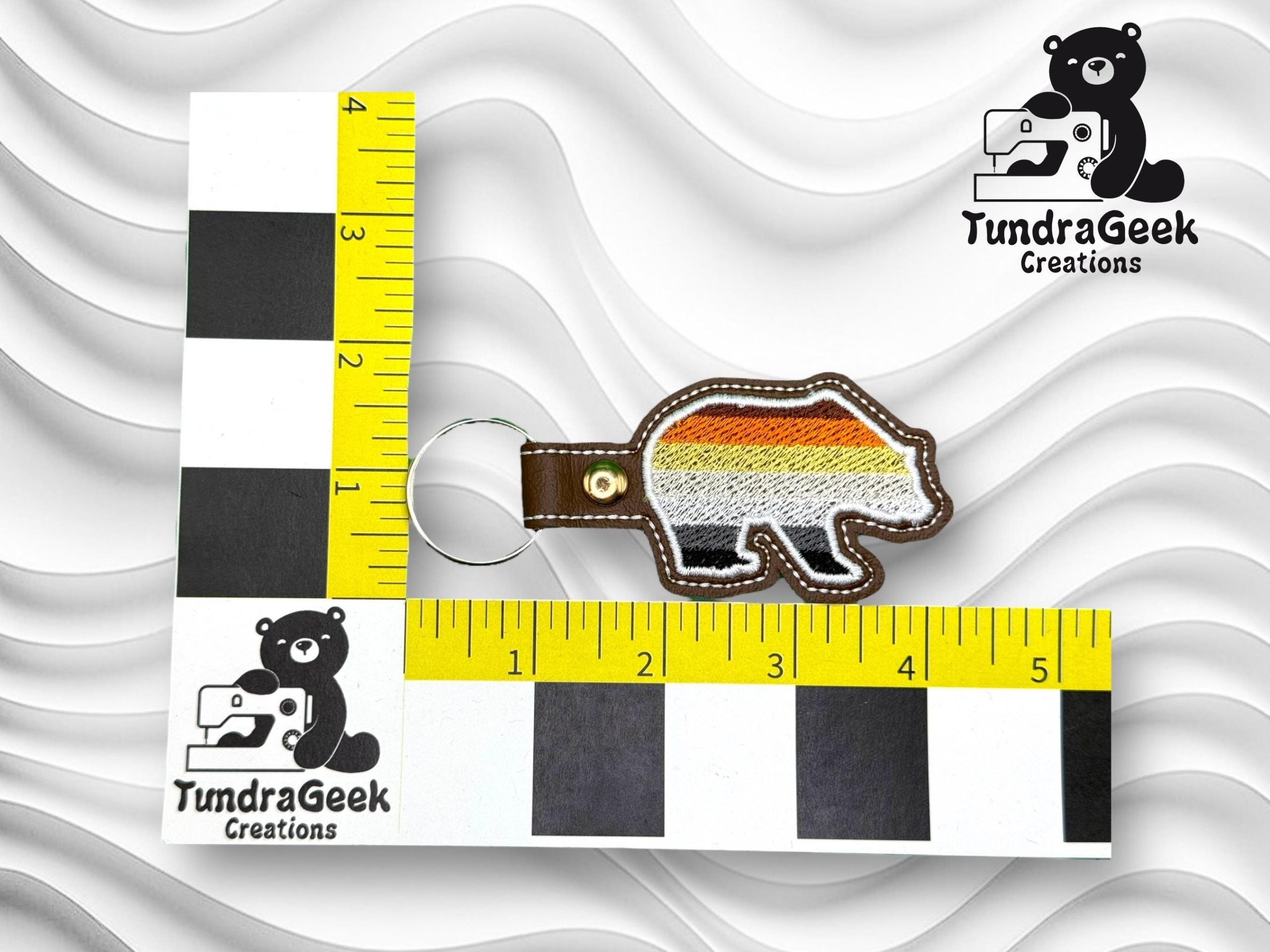 A bear-shaped keychain with a rainbow-striped design is shown next to two rulers for scale, featuring the TundraGeek Creations logo in two places.