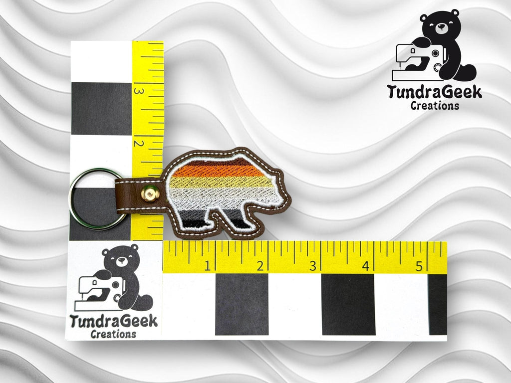 A bear-shaped keychain with multicolored stripes lies on a measuring grid showing it is about 3 inches long. TundraGeek Creations logos are visible in two corners.
