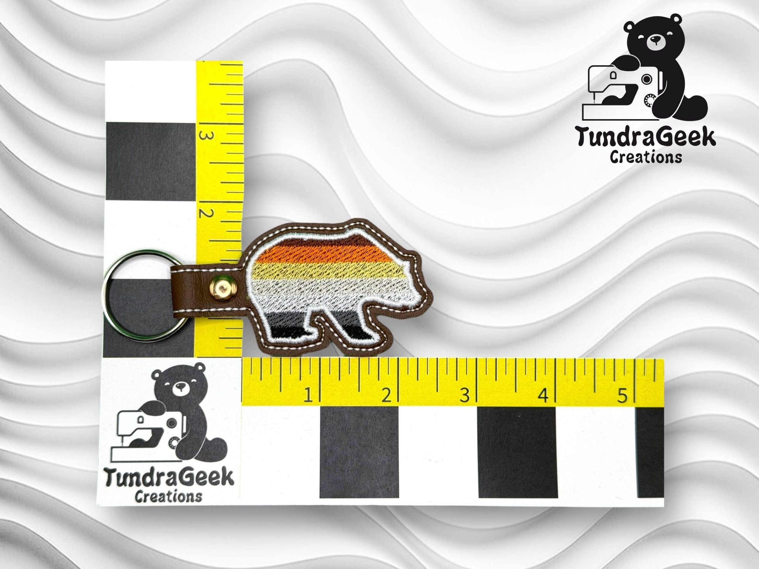 A bear-shaped keychain with multicolored stripes lies on a measuring grid showing it is about 3 inches long. TundraGeek Creations logos are visible in two corners.