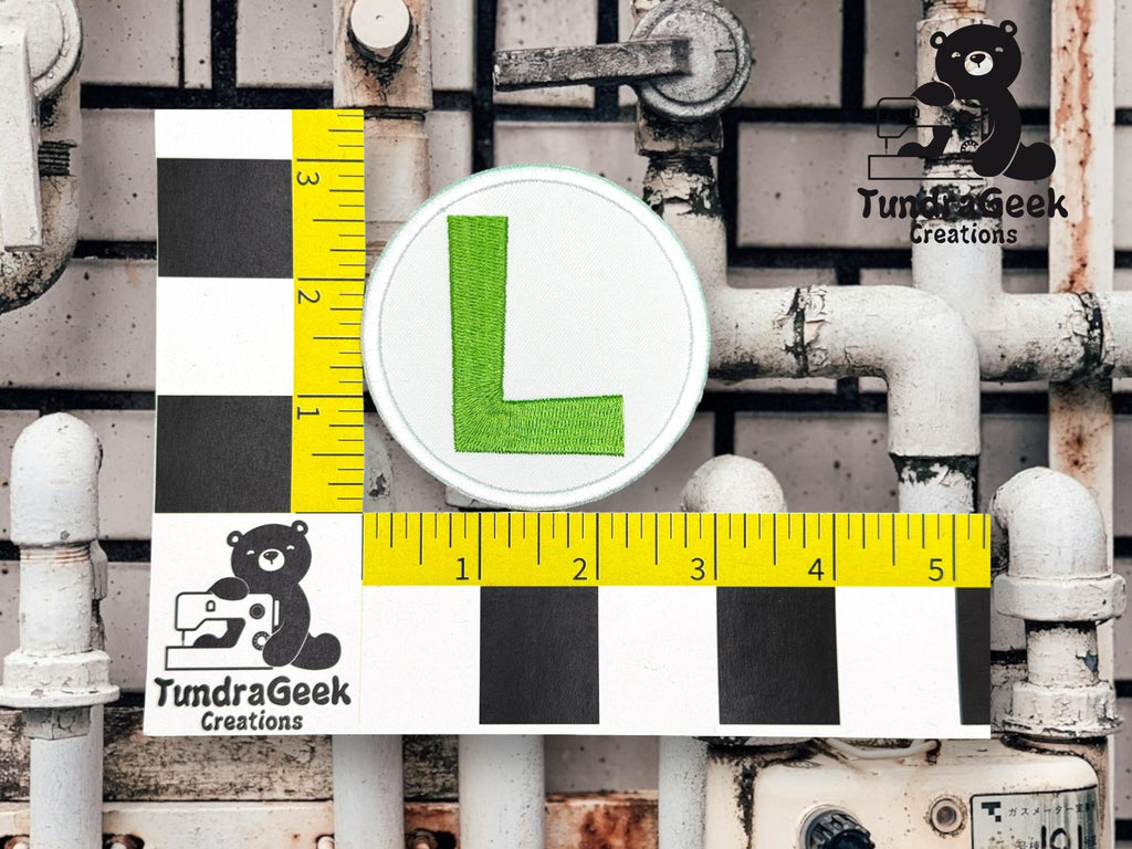 A round patch with a green L is measured next to a ruler and checkered scale, with TundraGeek Creations logos on a background of white pipes and a tiled wall.