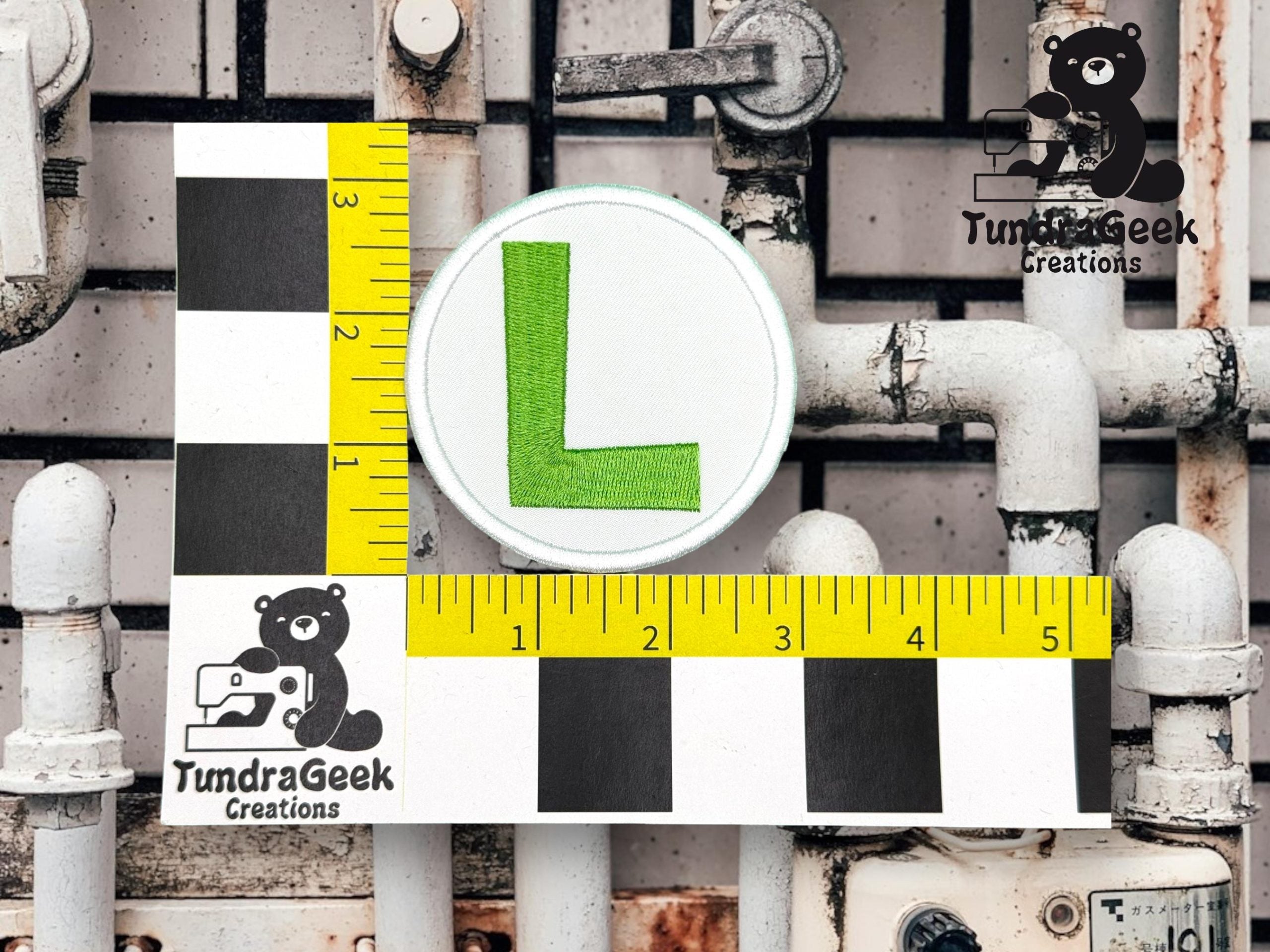 A round patch with a green L is measured next to a ruler and checkered scale, with TundraGeek Creations logos on a background of white pipes and a tiled wall.